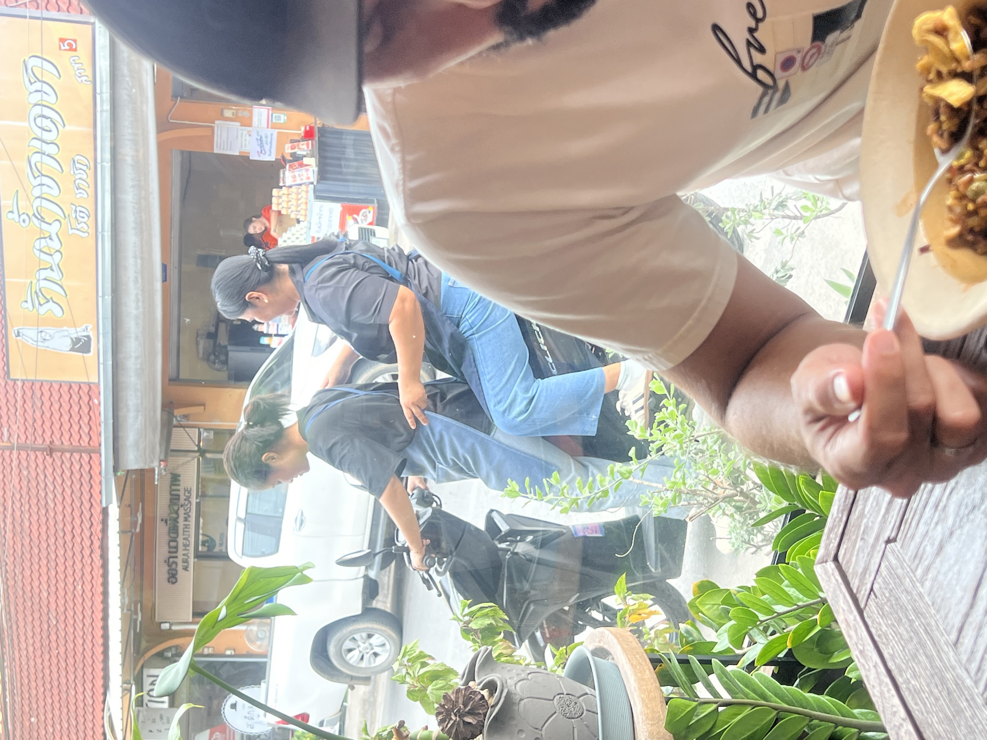 In an outdoor scene in Unknown, Unknown, two individuals are on a dark-colored motorcycle. The person in the front, wearing a black shirt and blue apron, is operating the vehicle. The person behind them, also in a black shirt and blue apron, is a passenger. They are on a paved surface, likely a street, with a white pickup truck visible behind them. In the foreground, a person's hands are visible, holding a fork over a bowl of food, suggesting a dining setting. Green plants are also present in the foreground, adding to the outdoor ambiance. The background shows a building with signage that appears to be in a foreign language, and several other people are visible inside the building. The lighting suggests it is daytime.