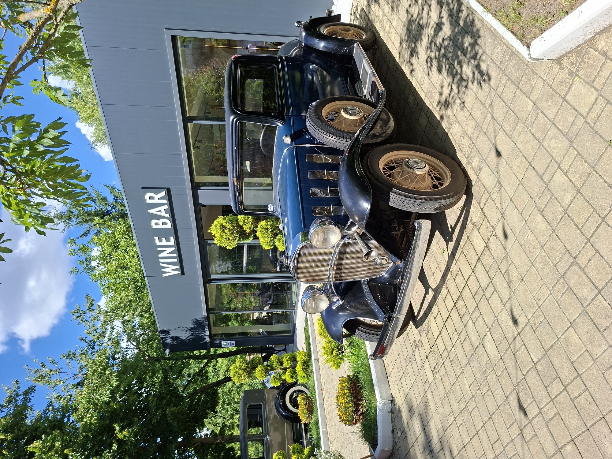 A dark blue vintage coupe automobile is parked on a light-colored paved area in front of a modern building. The building has large glass windows and a gray facade with the white text "WINE BAR" above the entrance. The car is positioned at an angle, facing towards the right of the frame, with its front grille and headlights prominently visible. The car features chrome accents, a spare tire mounted on the side, and light-colored wire-spoke wheels. Behind the vintage car, another older model car, a dark olive-green sedan, is visible parked further back and to the left. The environment surrounding the cars includes neatly trimmed green bushes and trees, with a blue sky and scattered white clouds overhead. The paved area is made of rectangular interlocking stones, with some shadows cast from the trees and the car. The scene appears to be an outdoor area, possibly a courtyard or entrance to a establishment.
