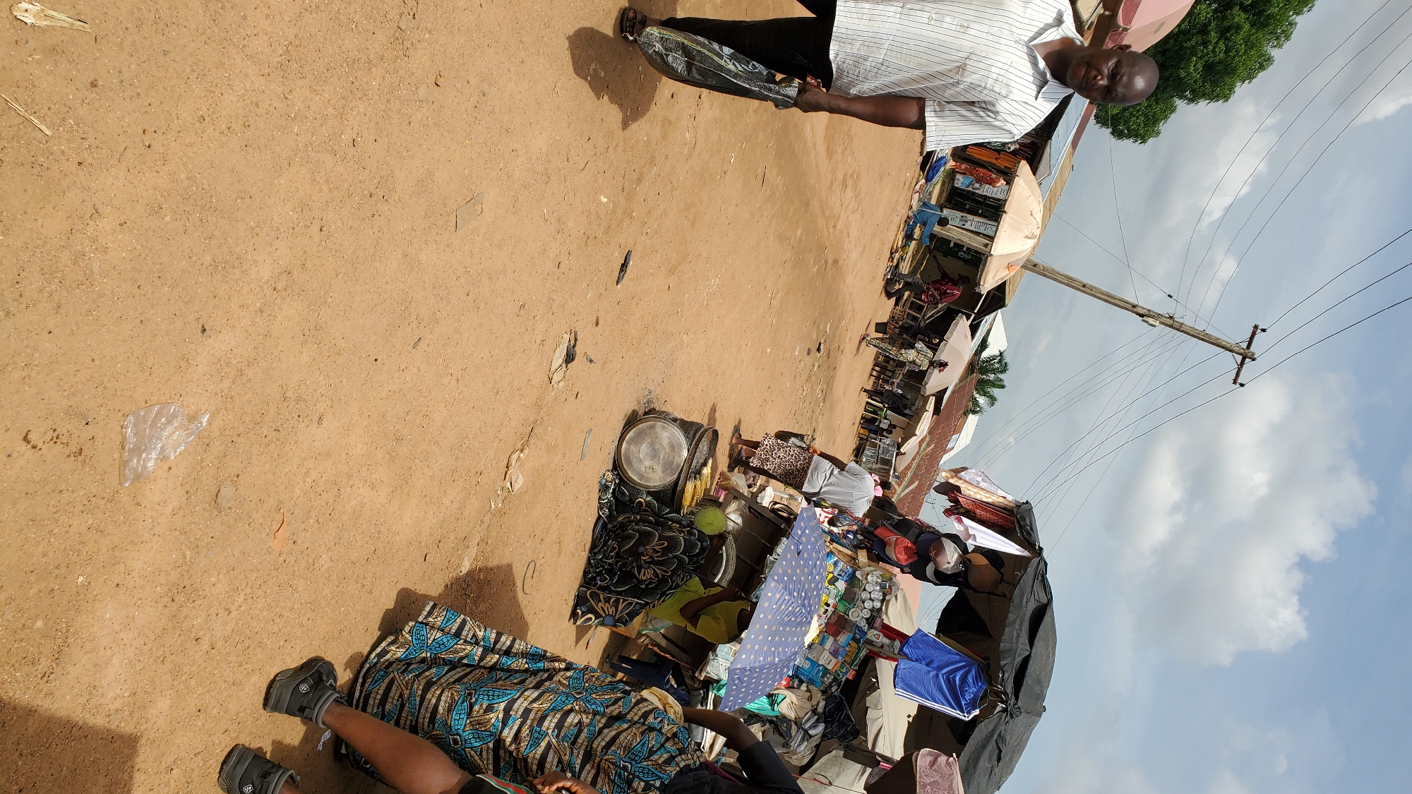 This is a medium-shot, eye-level, vertical, outdoor, daytime photograph taken in a market in Jalingo, Nigeria. The scene depicts a bustling marketplace with various stalls and people. The ground is sandy and dusty, typical of a rural or semi-urban environment.
In the foreground, a man wearing a striped white shirt and black trousers is walking towards the camera, carrying a long, dark, flat object. To the right of the man, a woman is partially visible from the waist down, wearing a colorful patterned skirt and black sneakers. She is standing near a stall where corn is being sold.
The middle ground of the image shows several market stalls with umbrellas and tarpaulins providing shade. Various goods are displayed, including clothing and food items like corn on the cob. People are seen browsing and interacting at the stalls. A prominent utility pole with electrical wires stretches across the background, suggesting the presence of electricity in the area. The sky is partially cloudy, indicating a typical weather condition.
The overall impression is one of a lively, everyday market scene in Jalingo, Nigeria, capturing the daily economic activities of the community.