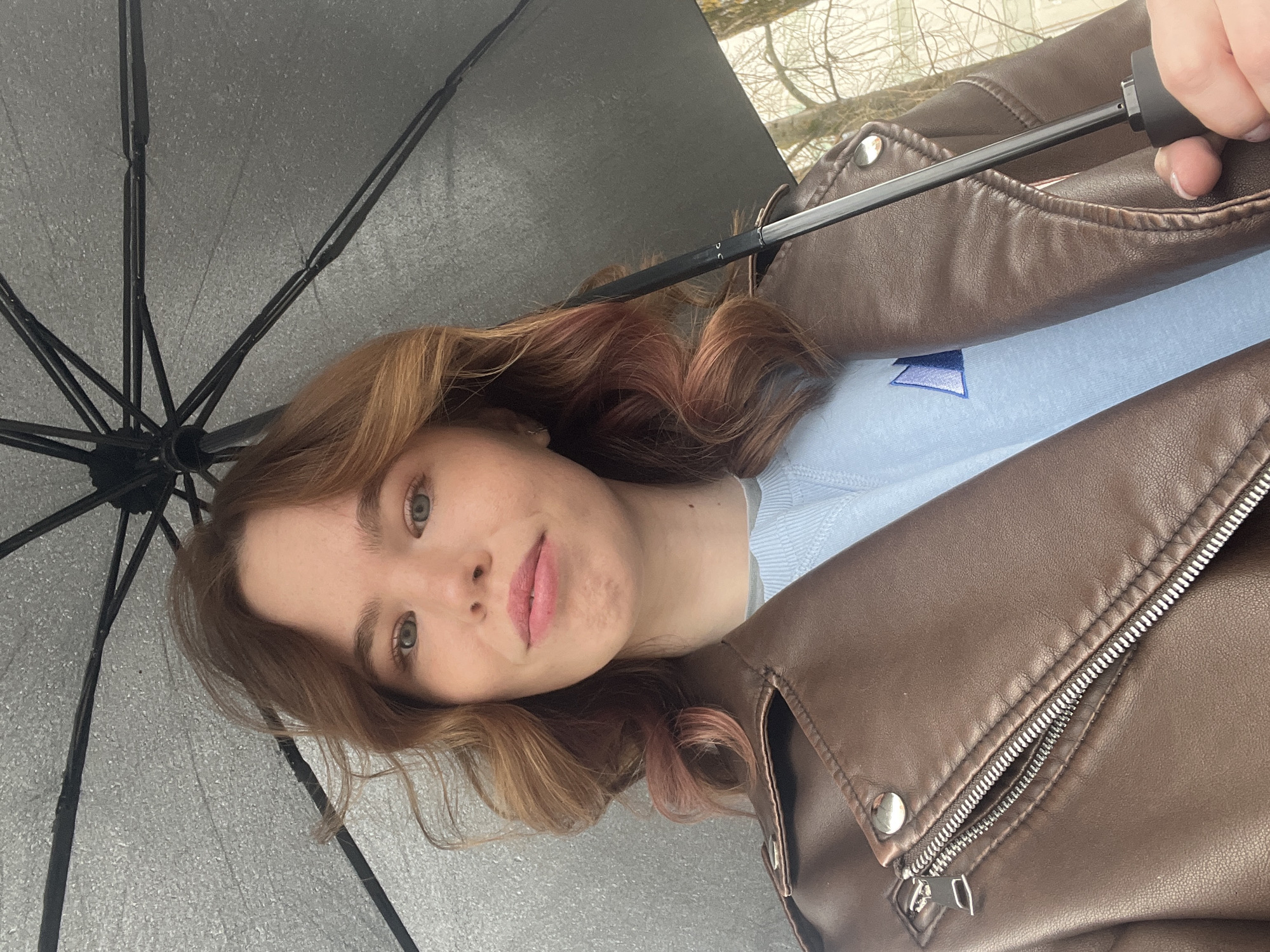 A young woman is holding an umbrella in an outdoor setting. She is wearing a light blue sweater and a brown leather jacket. The umbrella is open and provides cover from the elements. The background appears to show trees without leaves, suggesting it might be late autumn or winter. The location is unknown and can not be determined based on the image. The subject is holding the umbrella with one hand.