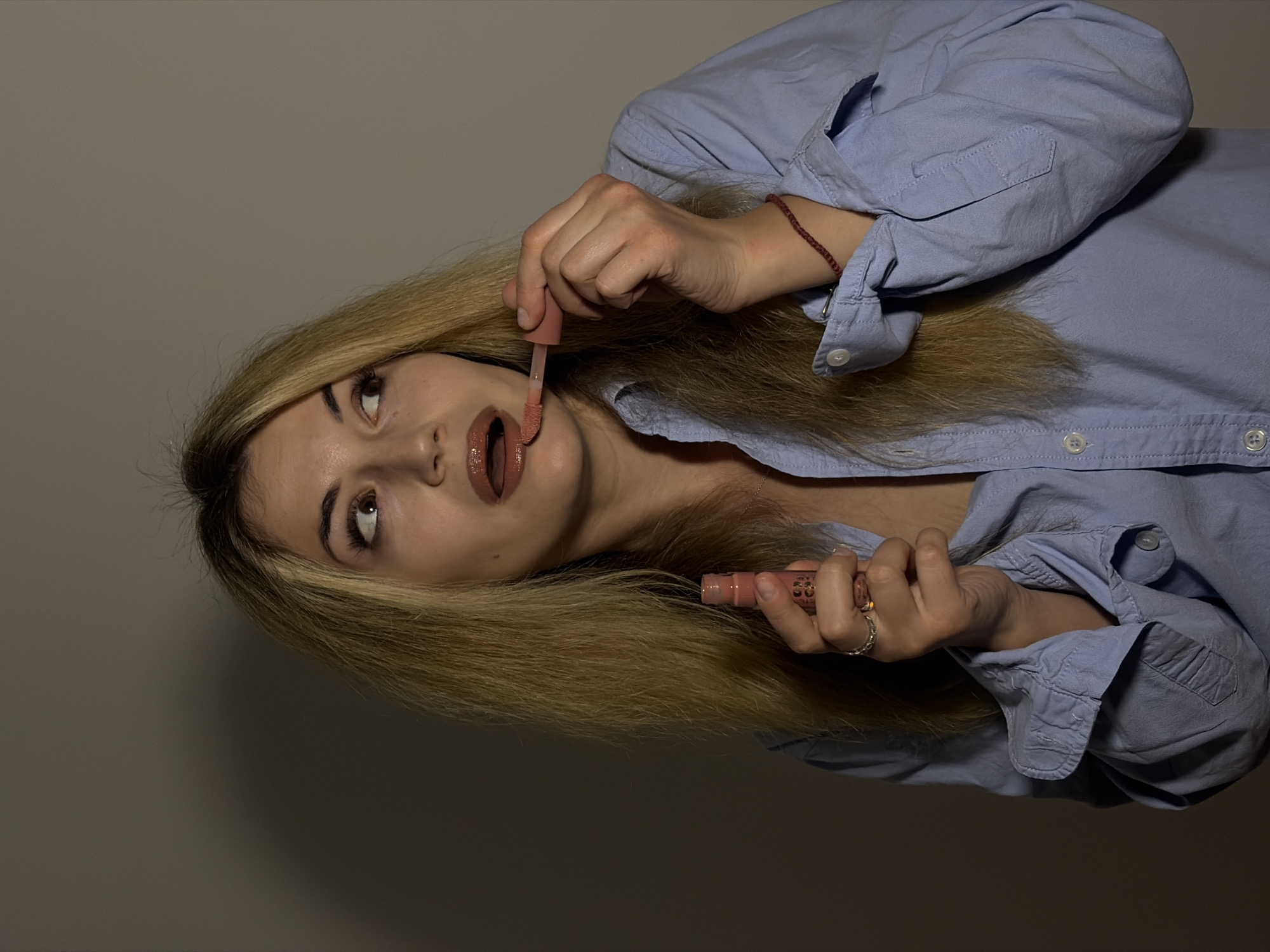 A young woman with long, straight blonde hair is captured in an indoor setting, applying lip gloss. The image is oriented with her head significantly tilted to the right. She has brown eyes, accentuated by dark eyebrows, and is looking upwards and to the right.
She is wearing a light blue, long-sleeved button-up shirt. A thin red string bracelet adorns her right wrist, and she has a silver-toned ring on her right ring finger. She holds a tube of peachy-nude or light brown liquid lip gloss in her left hand and uses the applicator, held in her right hand, to apply the product to her slightly open mouth.
The background is a plain, dark gray wall, indicating an unidentifiable indoor location. The lighting is somewhat dim, highlighting her face and upper body while leaving the background in shadow. While some text is faintly visible on the lip gloss tube, it is illegible. The scene depicts a moment of personal grooming, with the woman focused on the task. The location context provided is Budva, Montenegro, though no visual cues within the image confirm this.