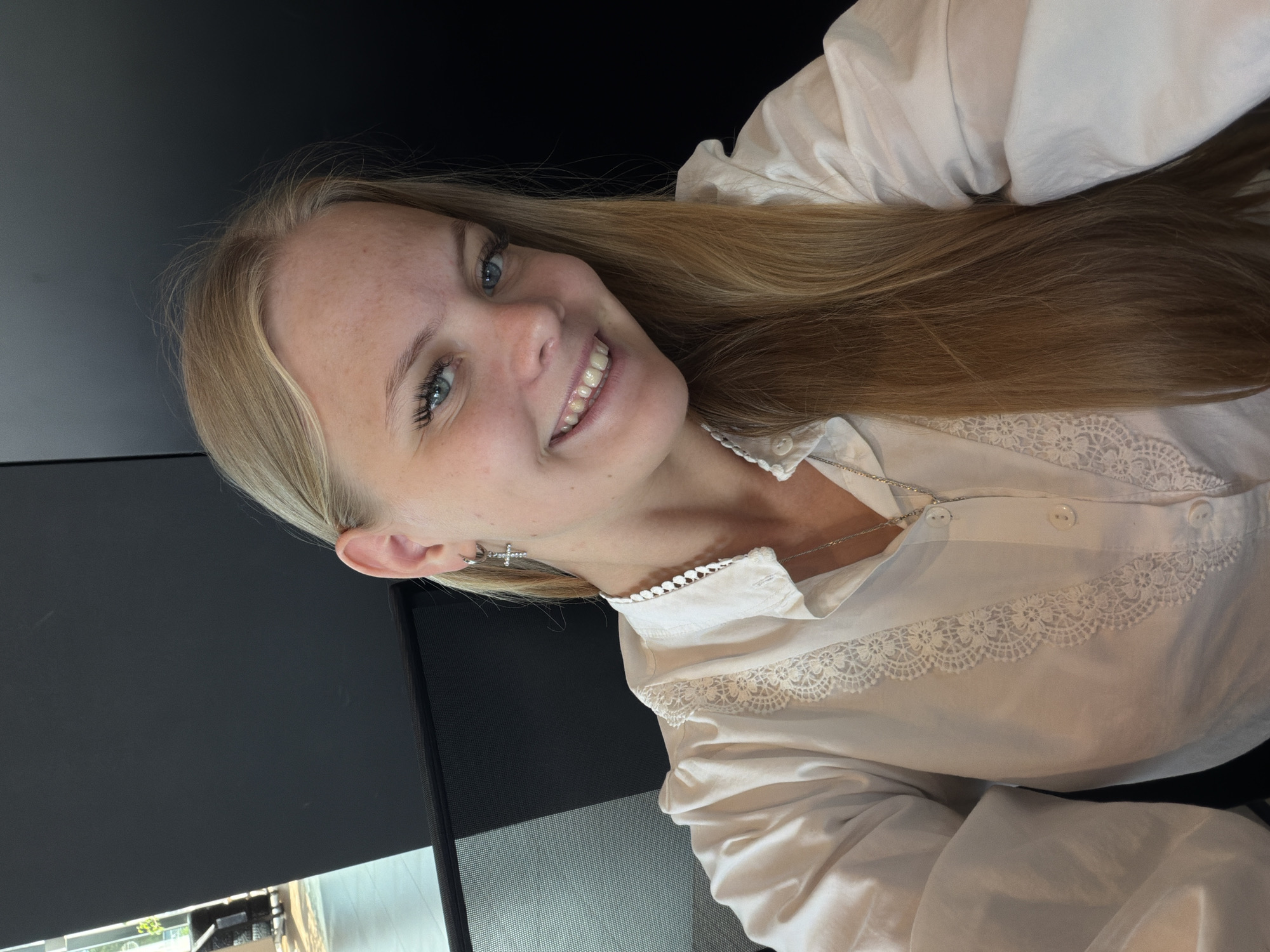 A young woman with blonde hair and blue eyes smiles into the camera. She is wearing a white collared shirt with lace detailing and a silver cross necklace. She has a silver hoop earring with a cross pendant in her left ear. The background is dark, with a hint of a modern office environment visible on the left. The image appears to be a selfie.