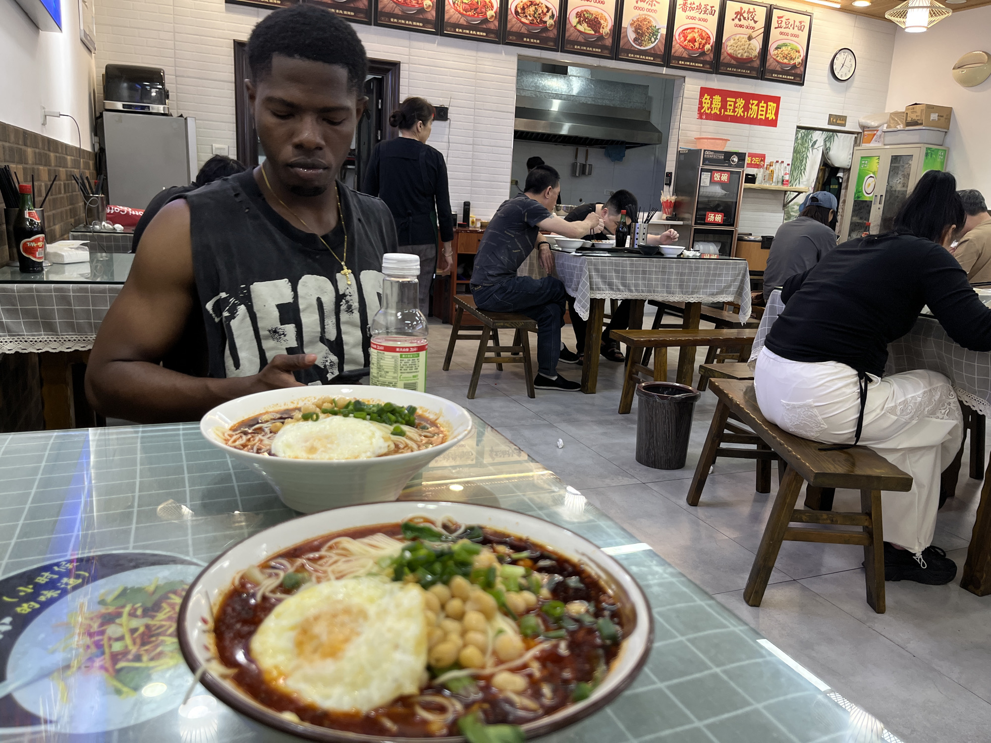 The photograph depicts the interior of a restaurant in Chongqing High-tech Zone, China. In the foreground, a Black man is seated at a table, looking down at his phone. Two bowls of noodles, each topped with a fried egg, are prominently displayed on the table. One bowl also contains chickpeas and green onions. A bottle of water is visible near the man.
In the background, several other patrons are seated at tables, eating. A woman in a black shirt and grey pants is walking toward the kitchen. There is a kitchen area with stainless steel equipment.
A menu board is visible above the kitchen area, displaying various noodle dishes with Chinese characters and phone numbers. A clock hangs on the wall, and a small refrigerator is present near the entrance. The tables are covered with a gray and white checked tablecloth. The restaurant has tiled walls and a tiled floor.