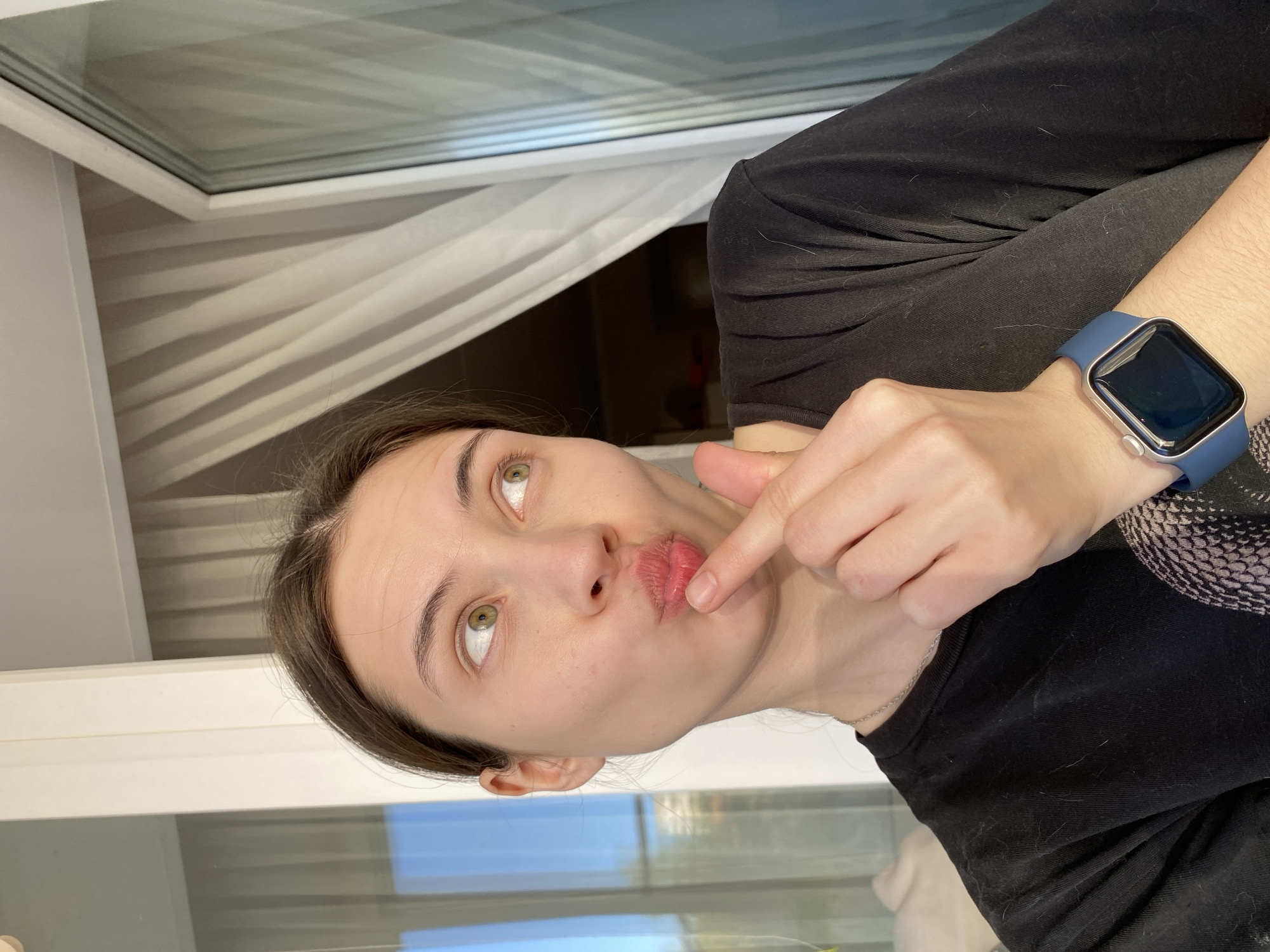 A young woman is indoors, possibly in Votkinsk, Russia. She is fair-skinned with brown hair, green eyes, and is wearing a black top. Her lips are pursed, and her right index finger is touching her bottom lip. A blue smartwatch is on her left wrist. A window with white frames and blinds is visible in the background.
