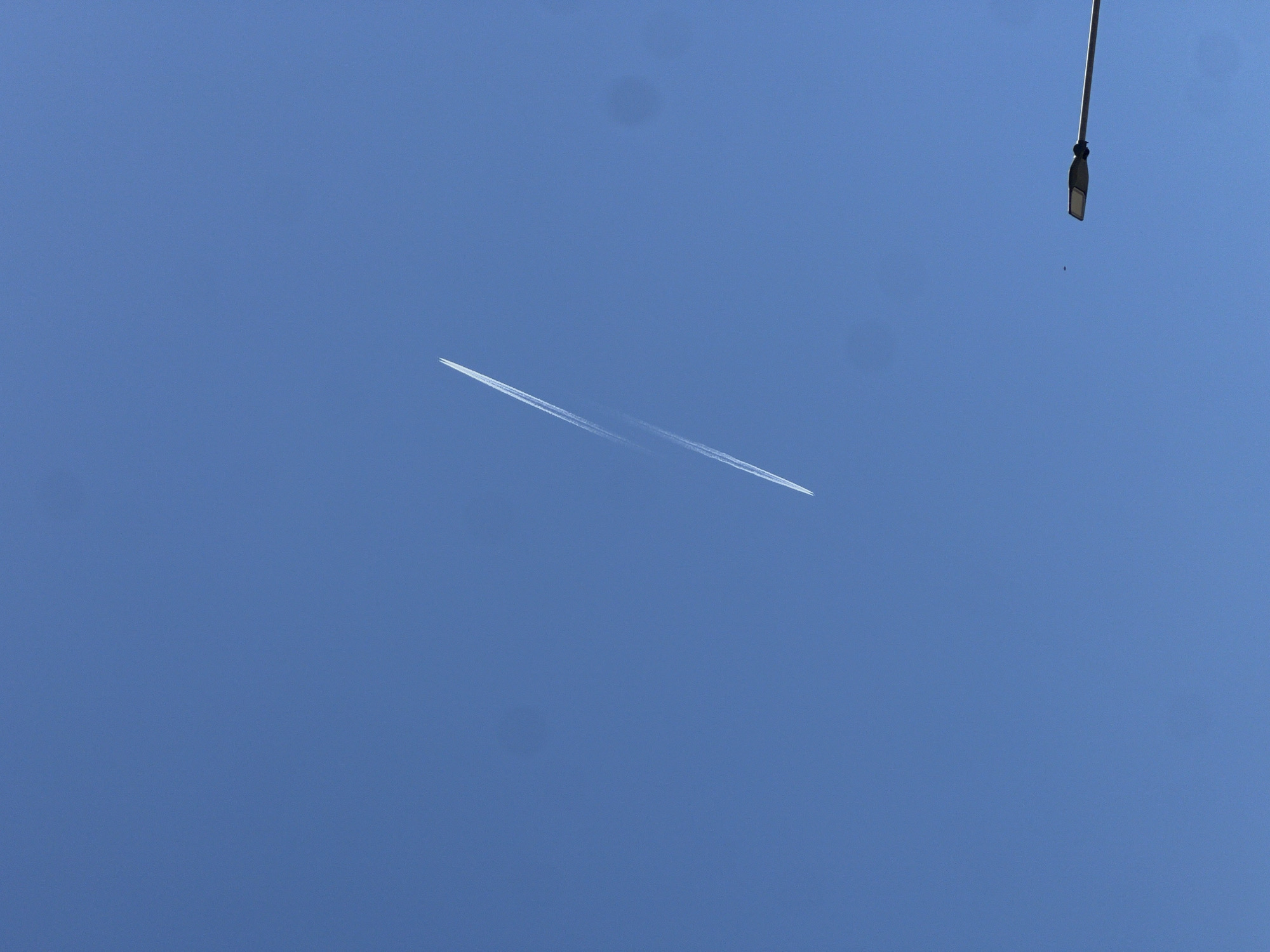 A photograph shows a clear, blue sky with two contrails intersecting near the center of the frame. In the upper right corner, there is a street lamp with a vertical pole. The location is implied to be the city of Bar, Montenegro, based on the prompt.