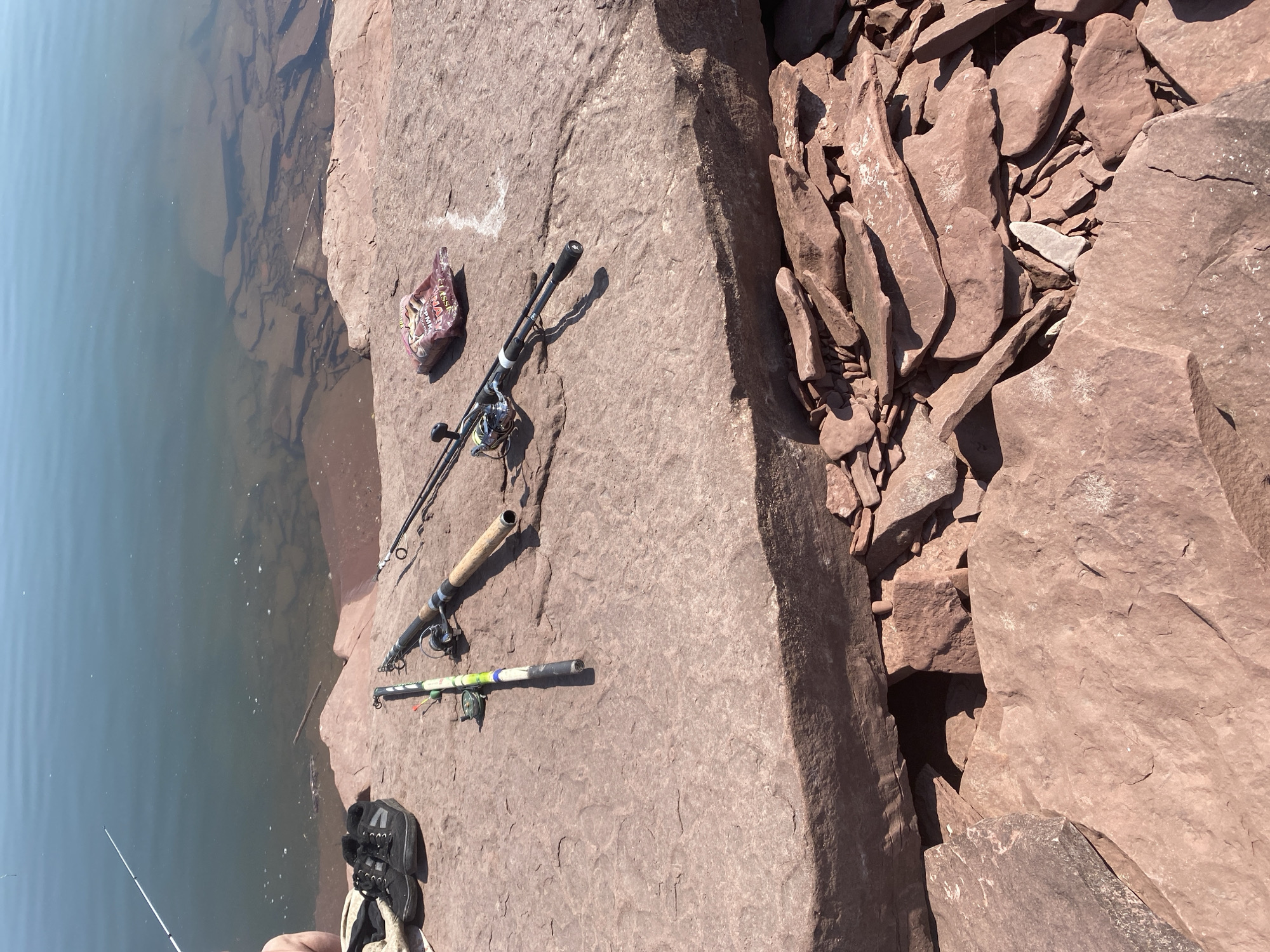 The image shows a rocky shoreline next to a body of water. On the rocks are two fishing rods, a pair of black sneakers, and a small bag of bait. One fishing rod is mostly yellow and green, while the other is black with a wooden handle. The sneakers are on the left side of the image, and the bait bag is to the right of the fishing rods. The water is calm and reflects the blue sky. The rocks are a reddish-brown color and appear to be sedimentary. There is a person's leg visible on the far left of the image, suggesting someone is fishing. The scene is set in the city of Ust'-Erba, Russia, known for its natural beauty and outdoor recreational opportunities.