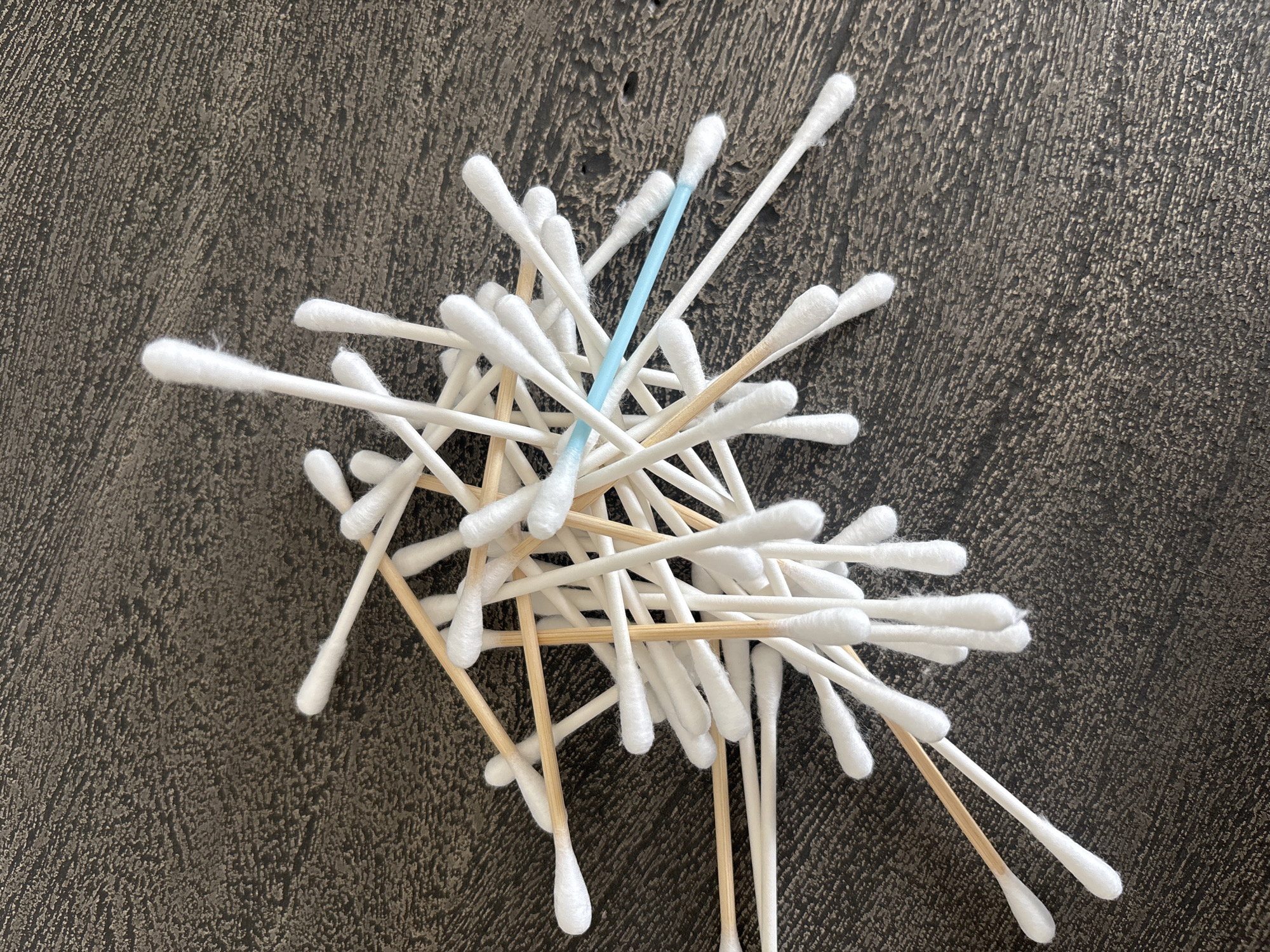 A pile of cotton swabs, some with white cotton ends and wooden handles, and one with a blue plastic handle, are scattered on a textured dark brown surface. The swabs are of various lengths and are jumbled together, with some overlapping others. The surface appears to be a tabletop or counter. There is no indication of the location in Dubai, United Arab Emirates.