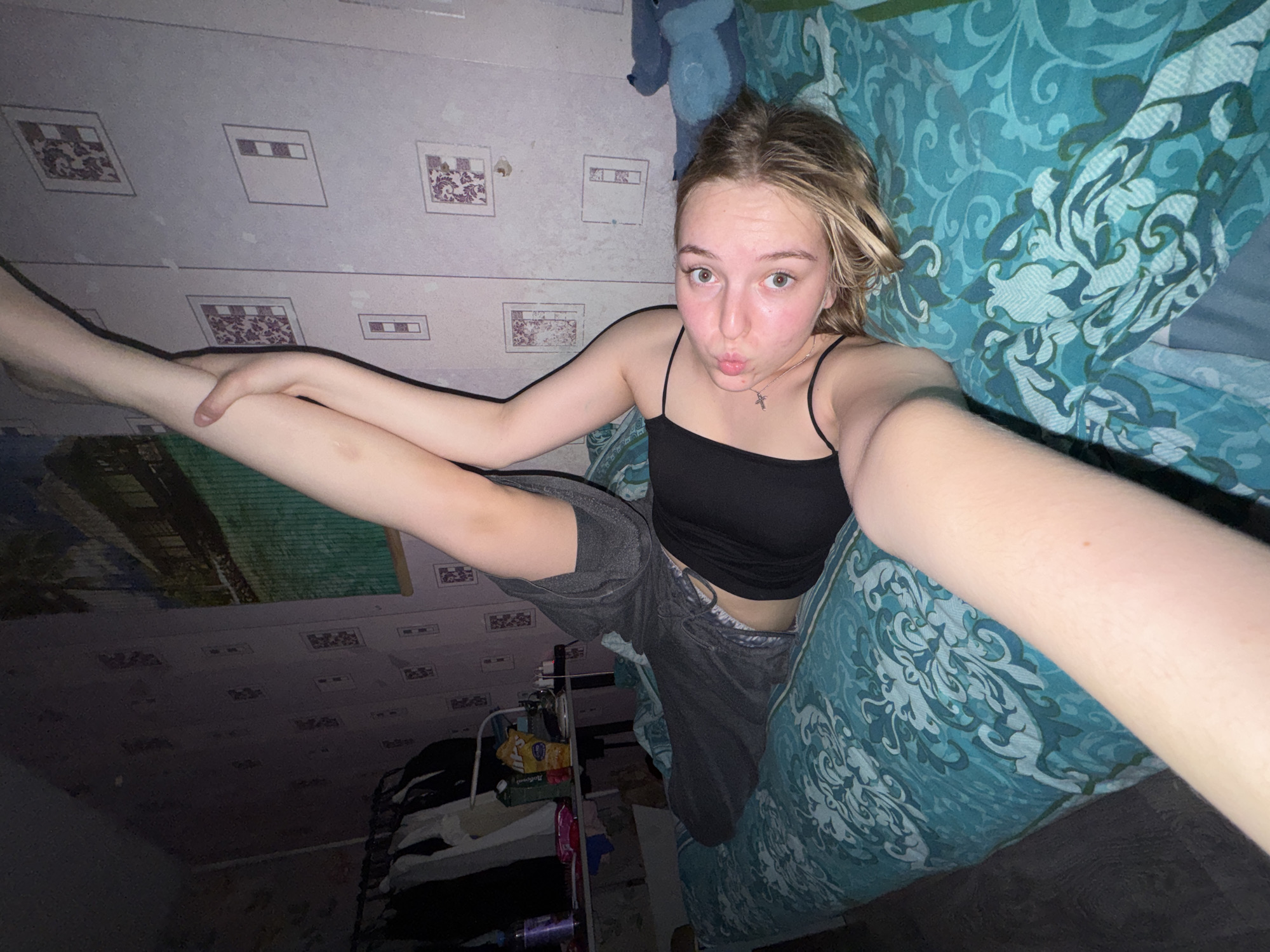The image appears to be a selfie taken indoors. A young Caucasian female with blonde hair is the central figure. She is wearing a black tank top and gray shorts or pants. She is holding one leg straight up in the air. She has a silver cross necklace. She is inside a room in Perm, Russia. The walls are light-colored with a repeating geometric pattern. The bed has a blue patterned bedspread. There is a stuffed blue animal on the bed. Below her is a shelf with a variety of items.
