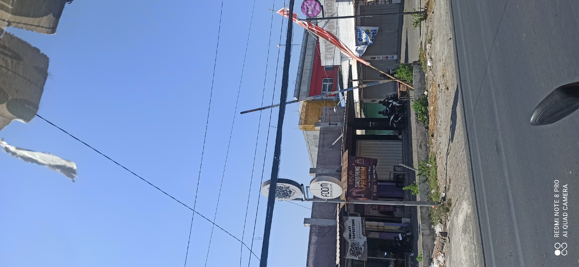 This is a ground-level, eye-level, daylight, outdoor, wide-angle shot of a street scene in Sedati Agung, Indonesia. The image captures a row of storefronts, a road, and a clear blue sky.
In the foreground, the asphalt road dominates the bottom half of the frame, with the edge of a dark-colored motorcycle seat visible at the bottom right. The sidewalk area in front of the storefronts is paved with concrete and uneven.
The storefronts appear to be single-story structures. The central shop has a white, corrugated metal roll-down door. To its left, a smaller storefront has a black and white sign that reads "MELATI VAPESTORE". Above the central shop, a circular sign with "FOOM" written on it, along with a logo, is mounted on a pole. Another circular sign, partially obscured, is positioned above the "FOOM" sign. To the right of the central shop, another storefront also has a white, corrugated metal roll-down door.
Above the storefronts, a network of black electrical wires crisscrosses the sky. There are also several poles supporting these wires and signs. A tattered white cloth or flag hangs from a wire in the upper portion of the image, caught by the wind.
Several objects and elements are visible:
* **People:** Two individuals are seated on motorcycles in front of the central storefront.
* **Vehicles:** Two motorcycles are parked on the sidewalk in front of the Melati Vape Store. Two more motorcycles are parked in front of the central storefront, with two people on one of them.
* **Signs:** Multiple signs are present, including "FOOM OFFICIAL SELLER", "MELATI VAPESTORE", a sign advertising "DISCA SALON SMOOTHING PREMIUM", and other indistinct signage. A purple circular sign with white text and a logo is visible on a pole to the far right. A red and white flag is attached to a pole on the right side of the image.
* **Environment:** The sky is clear and blue. The ground is paved with asphalt and concrete. Vegetation, including green bushes and dry grass, is growing in patches along the sidewalk and in front of the buildings. Debris and litter are scattered on the ground in front of the storefronts. A yellow wooden structure is visible on top of a building in the background. A red building with windows is also visible in the background.