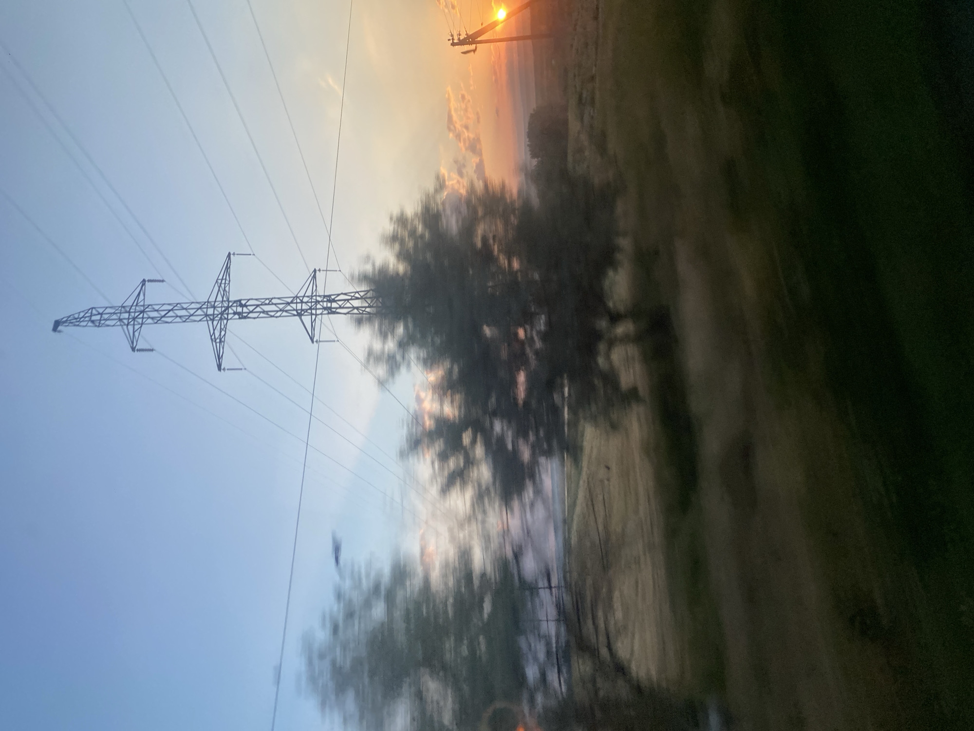 The image depicts a rural landscape at sunset, possibly in or near Novosiolovca, Moldova. A large metal electricity pylon dominates the upper center of the frame, with power lines stretching across the sky. In the foreground and midground, several trees are blurred due to motion, suggesting the image was taken from a moving vehicle. The setting sun casts a warm, orange glow across the sky, illuminating scattered clouds. A smaller electricity pole with wires is visible on the right side of the frame, with the sun directly behind it. The ground is covered in dry grass and fields. The overall impression is one of a serene, open landscape at dusk, marked by the presence of electrical infrastructure.