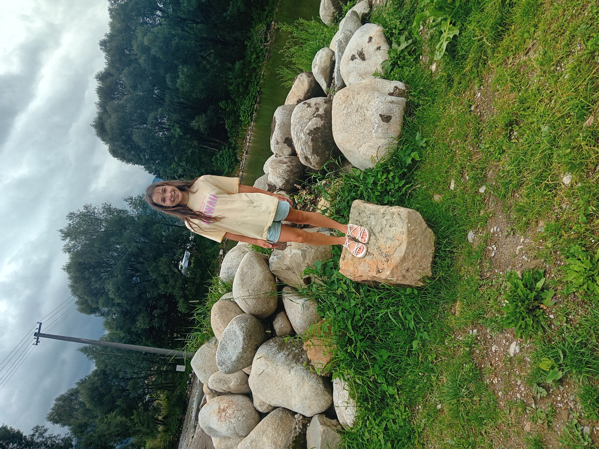 A young girl stands on a large rock in a natural setting, likely near Habovka, Slovakia, given the context. She is wearing a light-colored t-shirt with "HAWAII" printed on it in pink letters, and light blue denim shorts. Her feet are adorned with white sandals. The girl is smiling and looking at the camera. Behind her, a large pile of smooth, grey river stones is visible. Lush green grass and vegetation surround the stones and the rock the girl is standing on. In the background, a river with green water flows, bordered by a dense forest of dark green trees. The sky above is overcast with grey clouds. A telephone pole with wires is situated to the left of the frame, further in the background, adding to the rural or semi-rural feel of the location. The overall scene depicts a young person enjoying the outdoors in a scenic, natural environment.
