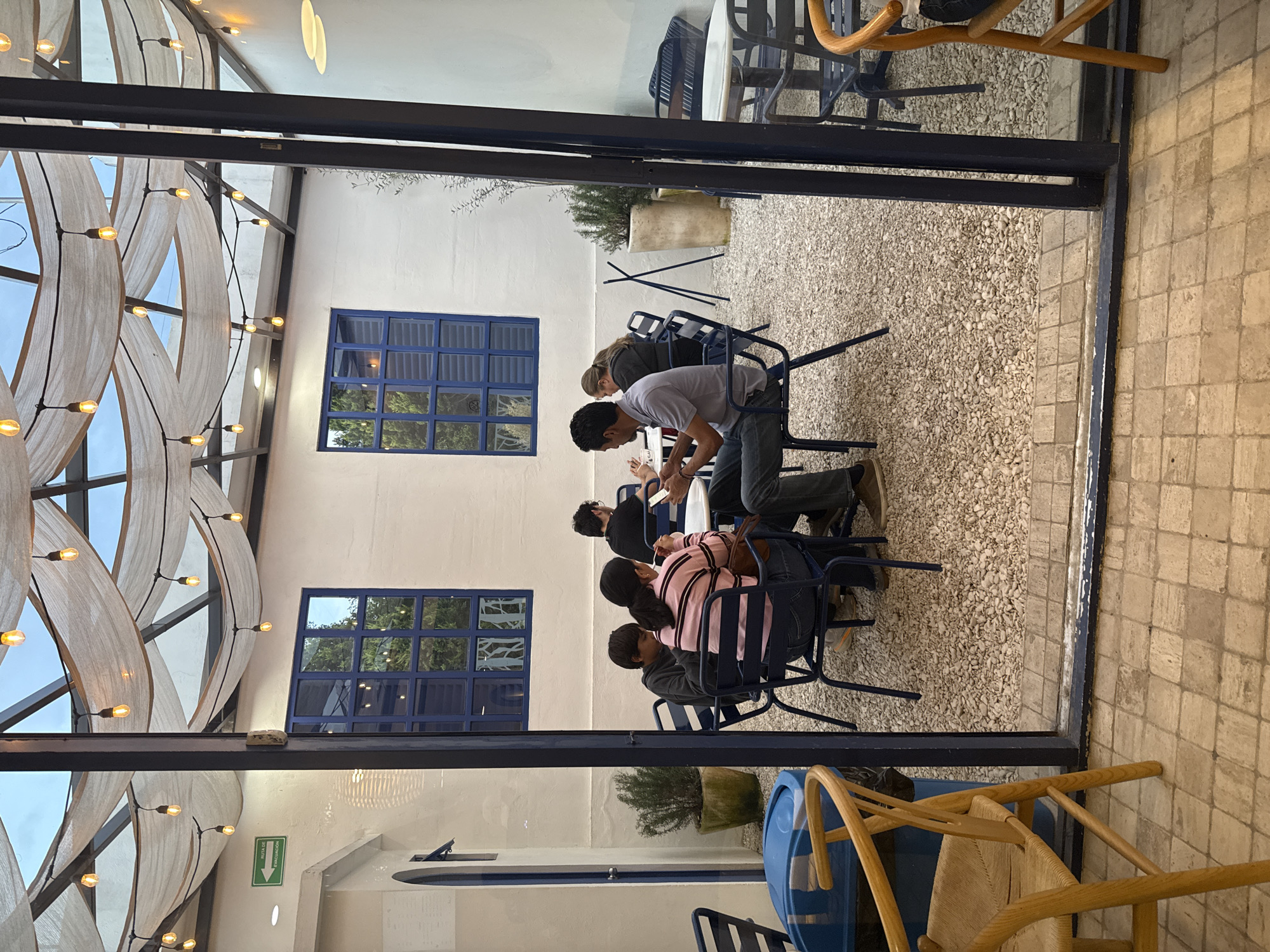 The image depicts the interior of a restaurant in Puebla, Mexico, viewed through a window or doorway. The restaurant has white walls with blue window frames. The flooring of the outdoor seating area is gravel. Several people are sitting at blue metal chairs around a table, seemingly engaged in a meeting. There is an emergency exit sign visible near the wall. The lighting is a combination of natural light and warm-toned decorative lights. There are plants in pots and other tables and chairs visible outside the main seating area.