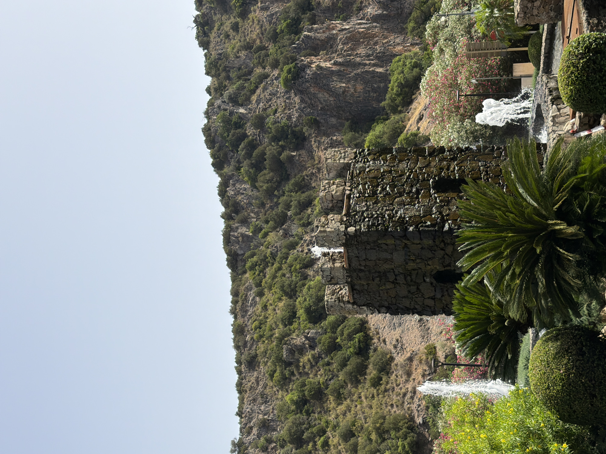 This is a medium shot, eye-level, outdoor photograph of a stone tower with water features and a rocky, vegetated hillside in the background. The tower is dark gray and constructed from irregular stones, with water cascading down its sides and spraying from its top. In front of the tower, to the left, is a tall, thin jet of water from a fountain. To the right of this, and partially obscured by the tower, is a smaller fountain with multiple jets. In the foreground, lush green foliage, including palm-like plants and a rounded bush, dominates the lower part of the frame. Behind the fountains, a hillside is covered in dense green trees and shrubs, with visible rock formations and a clear, pale blue sky above. The scene appears to be located in Benahavís, Spain, suggested by the architectural style and the Mediterranean vegetation. There are no people visible in the image.