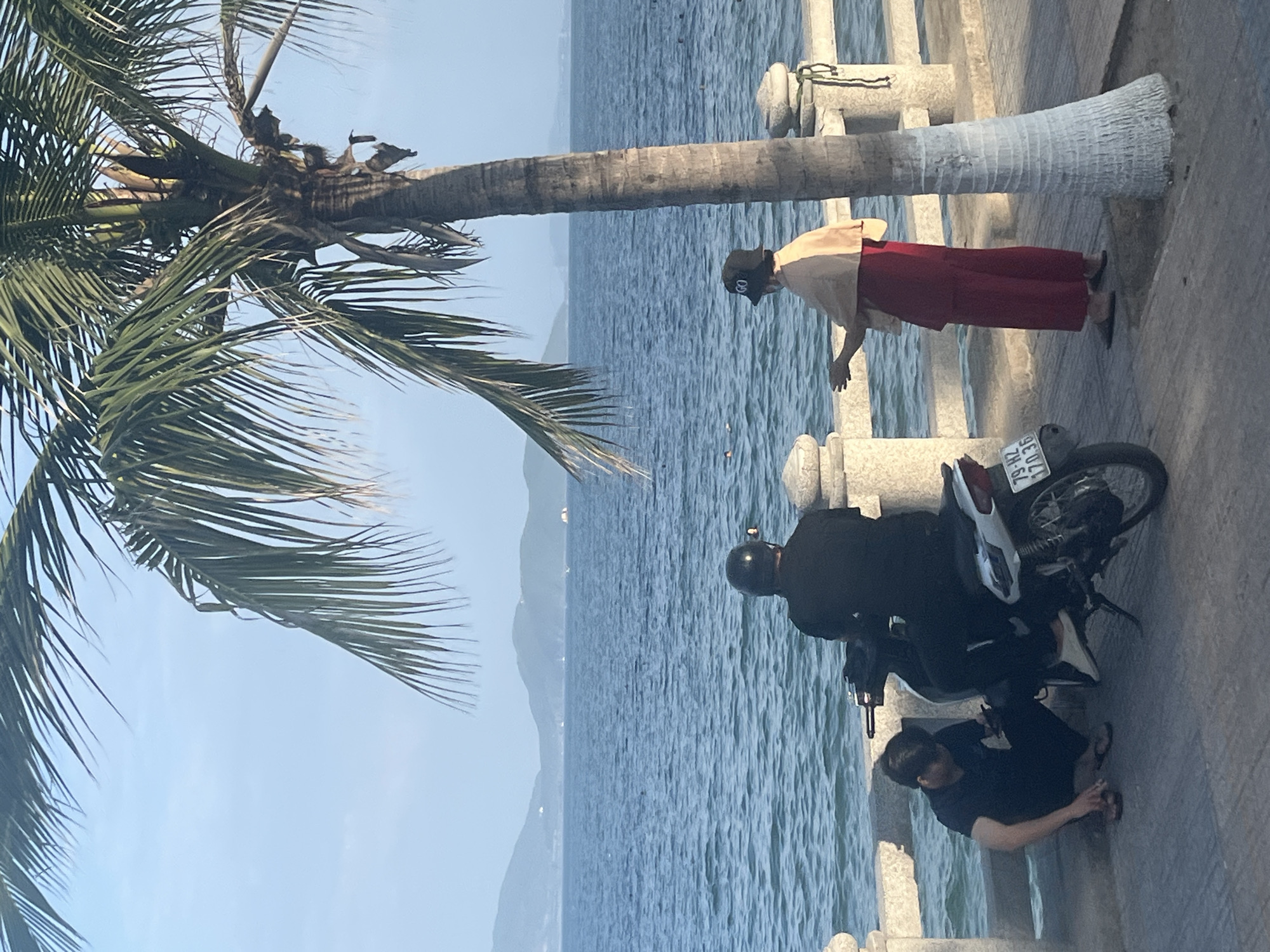 In Nha Trang, Vietnam, a man wearing a black helmet and black clothing sits on a motorcycle. Beside him, a man in a black t-shirt and black pants crouches, looking at his phone. To the right of the motorcycle, a person wearing a beige shawl over a red outfit and a black hat that reads "GO" stands by a palm tree. The individuals are positioned on a paved walkway next to a stone railing, with the ocean stretching out in the background. Distant islands and a clear sky complete the scene.