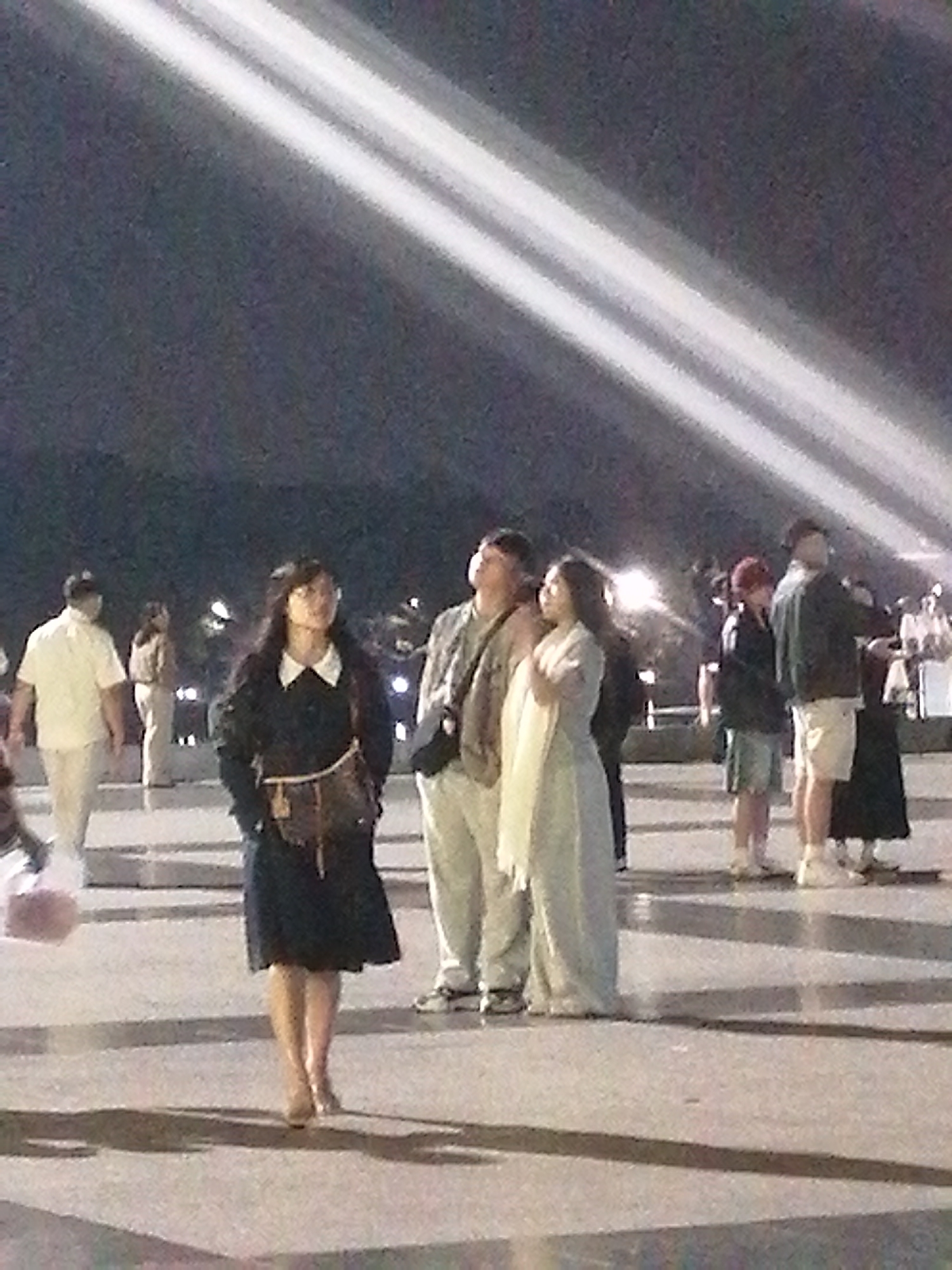 The image depicts a group of people standing in what appears to be an outdoor public space at night. The lighting suggests a performance or event, with strong beams of light cutting across the scene from above, illuminating the dark sky. In the foreground, a woman in a dark dress with a white collar is standing and looking slightly upwards. She has a bag slung across her body. Behind her, a group of people are gathered. To the left, a man in a white shirt and light-colored pants walks away from the camera. To the right of the woman in the foreground, a man in a tan jacket and light pants stands next to a woman in a light-colored dress. Further back, more people are visible, some in casual attire. The ground appears to be paved, with shadows cast from the people and the lighting.
While the image's content strongly suggests an event or gathering, there is no specific visual information within the image itself to confirm its location in Da Lat, Vietnam.