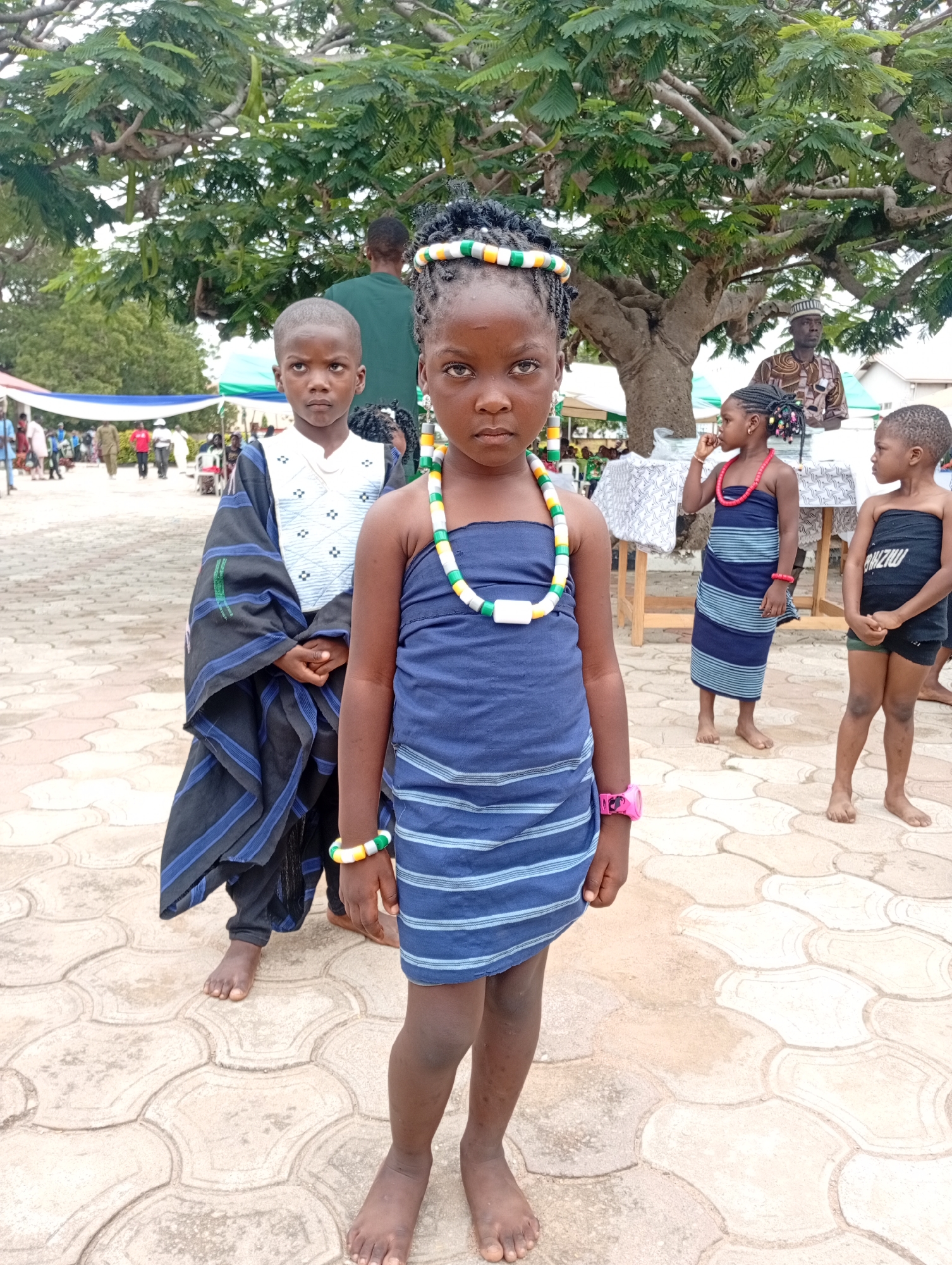In Barden Yana, Nigeria, a group of children in traditional attire stand on a patterned paved area, likely a courtyard or event space. The central focus is a young girl in a blue and white striped dress, adorned with a colorful beaded necklace and a matching bracelet. Her hair is styled in braids and decorated with beads. To her left, a boy wears a dark blue and black striped garment over a white shirt. Further to the right, another girl is dressed in a similar blue and white striped garment, with a red beaded necklace. A third child, on the far right, wears a black bandeau and shorts. In the background, adults are visible, some wearing traditional clothing. Lush green trees with thick branches provide shade over the scene. Several white and colored tents or canopies are set up in the distance, suggesting an outdoor gathering or festival. The ground is a light-colored, textured pavement with a repeating circular pattern.