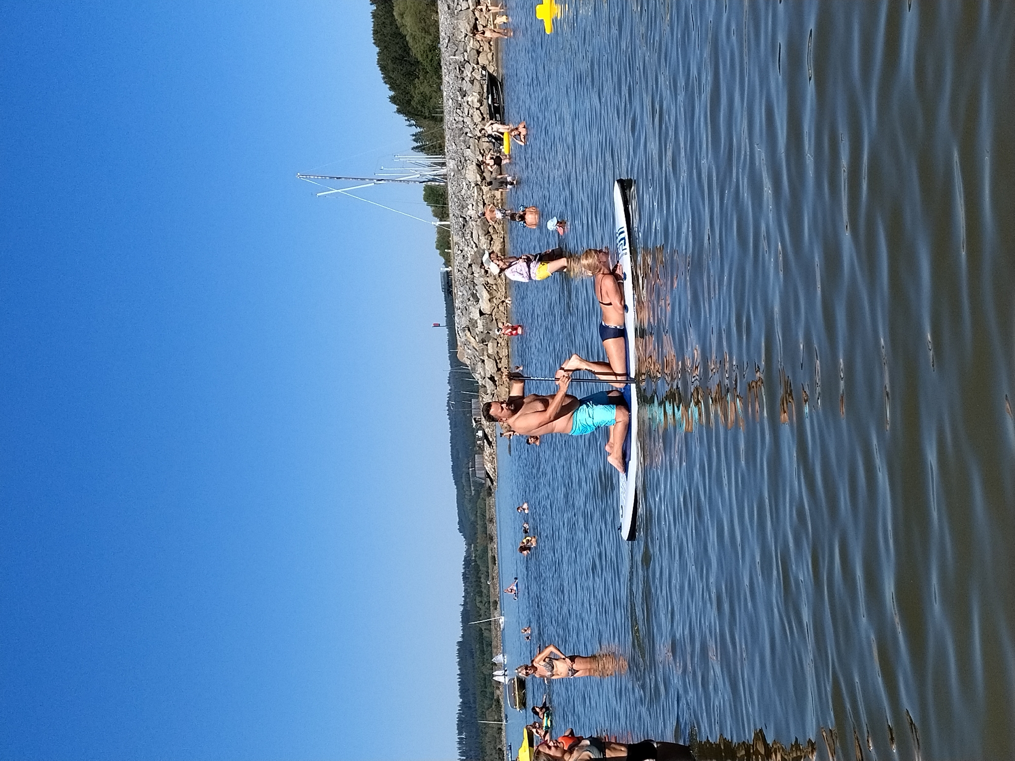 In Radslav, Czechia, on a bright, sunny day, a man on a paddleboard kneels in the water, holding a paddle. Another person lies prone on the same paddleboard, with their arms outstretched. They are surrounded by other people swimming and paddleboarding in the clear blue water of a lake. In the background, a rocky shore is visible, with trees and buildings beyond. Several sailboats are anchored in the distance. The sky is a clear, cloudless blue.