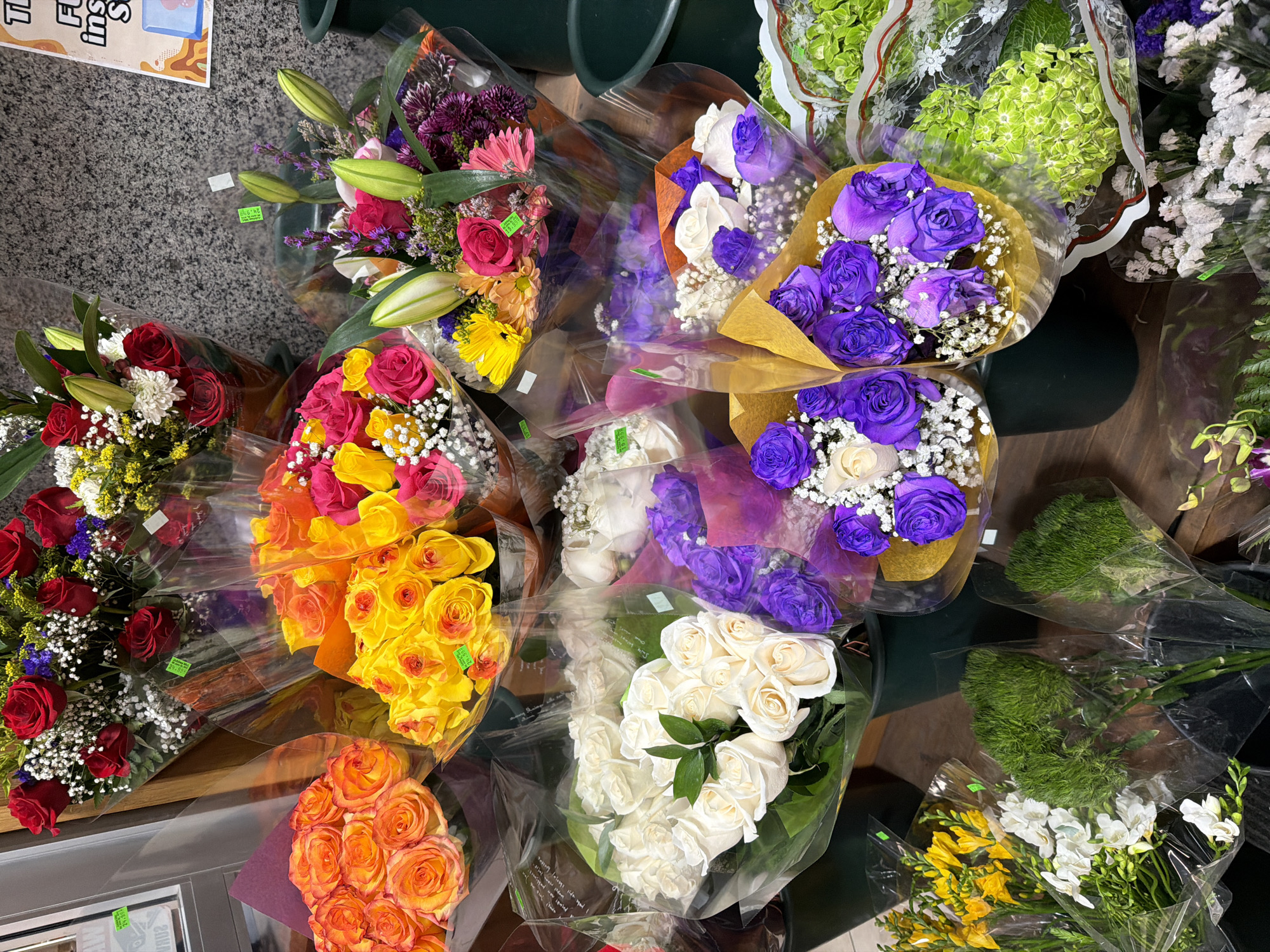 This is a medium shot photograph of a vibrant display of assorted flower bouquets. The flowers are arranged in multiple clear plastic wrappers and are presented at various heights and angles, suggesting they are for sale.
In the foreground, on the left, a bouquet of yellow freesia and white flowers is visible. To its right, two bouquets of bright green, fluffy flowers, possibly carnations or similar, are displayed in black plastic buckets. Further to the right, a bouquet of light green hydrangeas wrapped in clear plastic is prominent.
In the middle ground, a large bouquet of white roses is centered, with a bouquet of purple roses to its right, both wrapped in clear plastic with some colorful paper accents. Behind these, a bouquet of mixed yellow, orange, and pink roses and daisies is visible. At the top, a bouquet of red roses is featured.
The background consists of a textured, grey, speckled wall, reminiscent of granite or stone, which appears to be the interior of a store or market. A wooden element is visible on the left edge of the frame, possibly part of a shelving unit or display. A sign with some text and graphics is partially visible on the far right background.
The lighting is bright and even, illuminating the colors and textures of the flowers. No people are present in the image. The overall impression is a well-stocked floral display, likely in a flower shop or a market stall within New York City, USA.