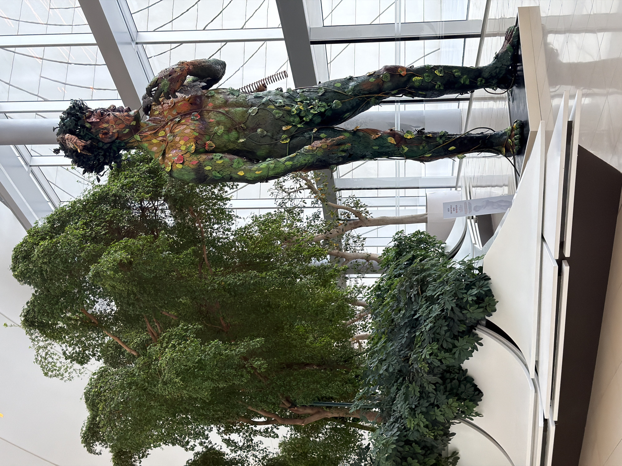 The image depicts a sculpture of a human figure covered in green vines and leaves, standing next to a large artificial tree. The sculpture is detailed, with the figure appearing to be made of a dark material, possibly metal or resin, and adorned with various colored leaves and branches. The artificial tree is lush and green, with a thick canopy of leaves. In the background, a modern building with large windows is visible, suggesting an indoor public space. The flooring appears to be polished tile or stone. The sculpture is positioned on a dark rectangular base. Several white, curved benches are arranged in the foreground. This scene is located in the Heydar Aliyev International Airport in Baku, Azerbaijan, where the "Spirit of Nakhchivan" sculpture by the contemporary artist Faig Ahmed is displayed.