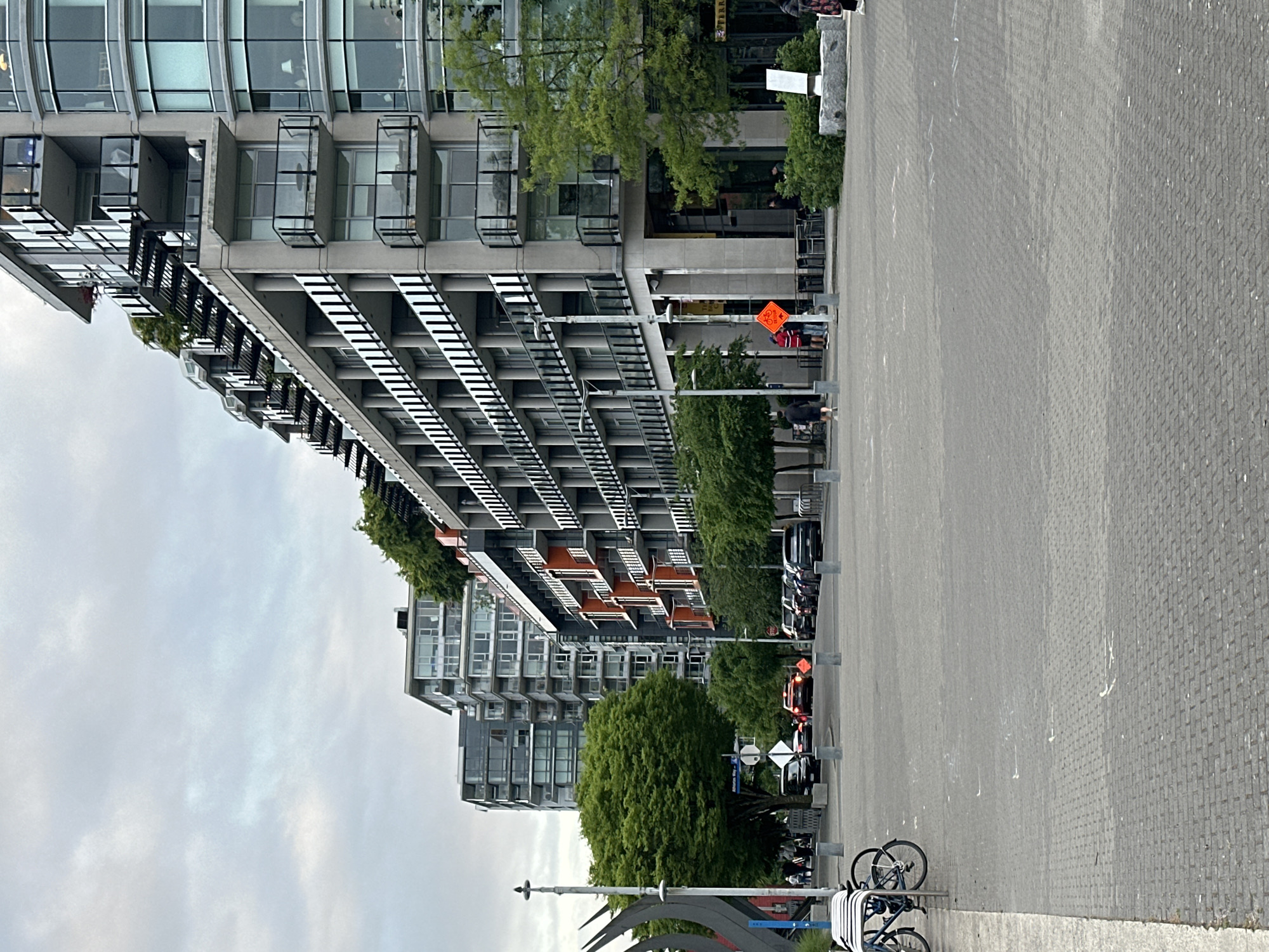 The image depicts a street scene in Vancouver, Canada. In the foreground, a paved walkway with a bicycle rack and parked bicycles is visible. The midground shows a street with parked cars and a pedestrian crossing. The background showcases modern architecture with multiple-story buildings featuring balconies and a mix of glass and concrete facades. Trees are interspersed along the street, providing greenery. Street signs and temporary construction signs are present, suggesting ongoing activity or maintenance in the area. The sky is overcast, contributing to a muted color palette.