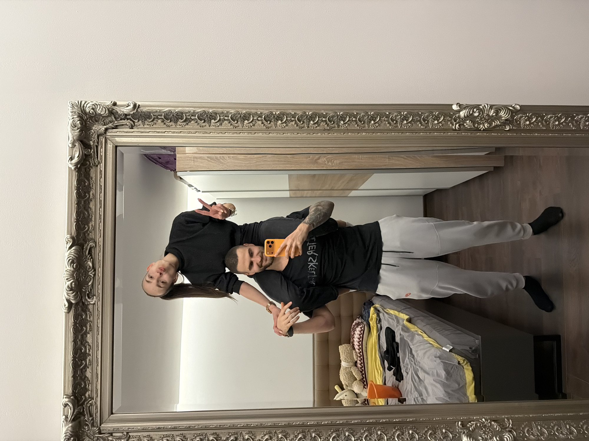 The scene depicts a mirror selfie taken indoors in a room in Sofia, Bulgaria. A male and a female, both appearing to be in their 20s or early 30s, are captured in the reflection of a large, ornate silver-framed mirror.
The male, with short dark hair and a beard, is standing, carrying the female on his shoulders. He wears a black t-shirt with a reversed white graphic, light grey sweatpants, and dark socks. Visible tattoos cover his left arm. He holds an orange-cased smartphone in his right hand, angled towards the mirror, indicating he is taking the photograph. His left hand supports the female's leg. He exhibits a slight smile.
The female, positioned on the male's shoulders, has dark hair tied in a high ponytail. She wears a black long-sleeved top and black pants. Her head is tilted to the left, and her lips are pursed in a "fish face" expression. Her right hand is raised, displaying a "peace sign." A gold-colored wristwatch is visible on her left wrist.
The background reflected in the mirror reveals a domestic setting. To the left, a bed with a brown headboard, grey and yellow bedding, and various stuffed animals (a white one and a beige teddy bear) is visible. A patterned pillow rests on the bed. An orange bucket and black fabric items are on the floor or bed surface near the foot of the bed. To the right, a white wardrobe with light wood paneling is present. The room has white walls and dark wood flooring.