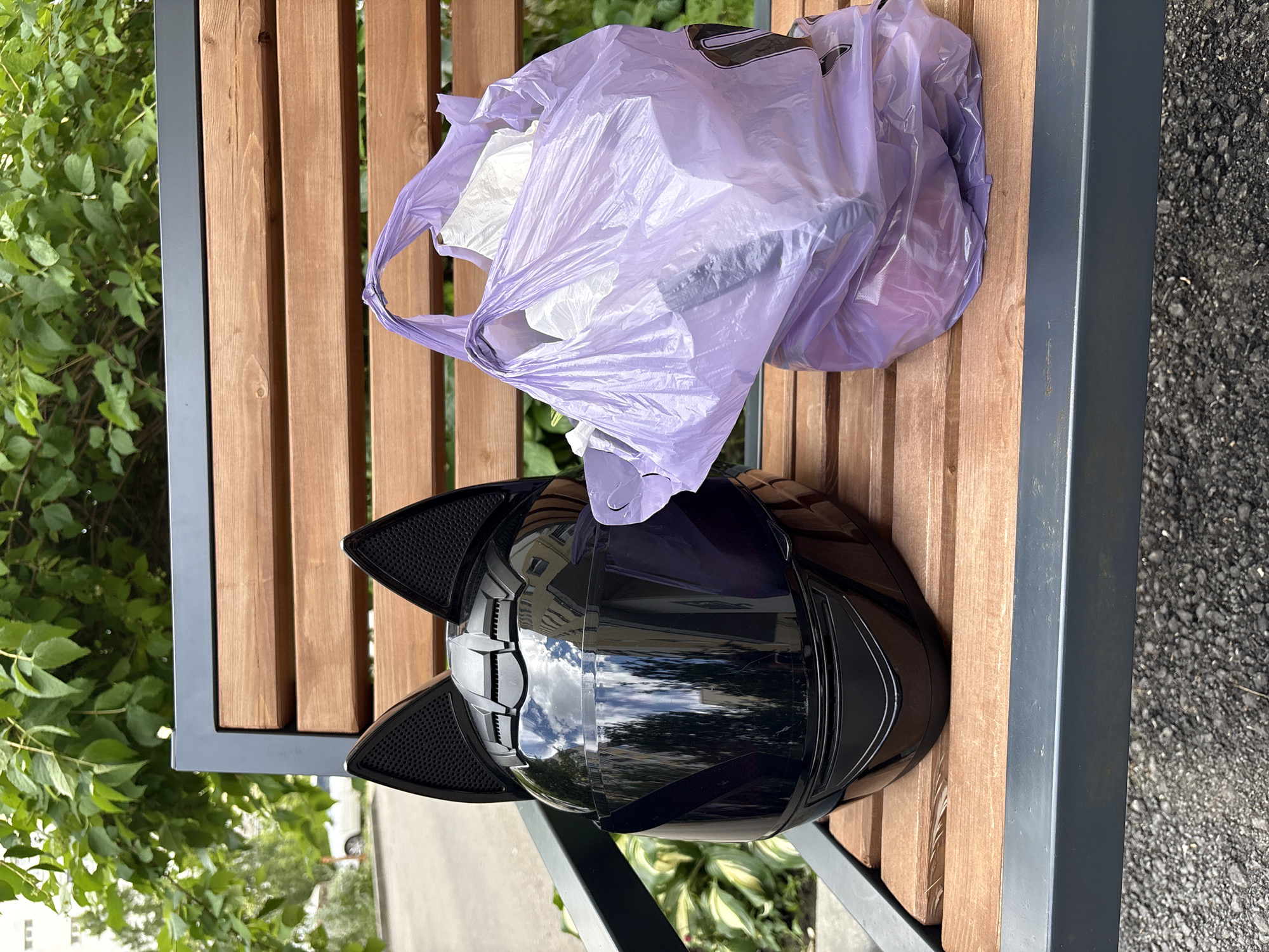A black motorcycle helmet with cat ears and a purple plastic bag filled with items are resting on a wooden park bench. The bench is situated outdoors, with lush green foliage visible in the background, suggesting a park or outdoor public space. Sunlight reflects off the helmet's visor. The scene is likely in Saratov, Russia, given the prompt.