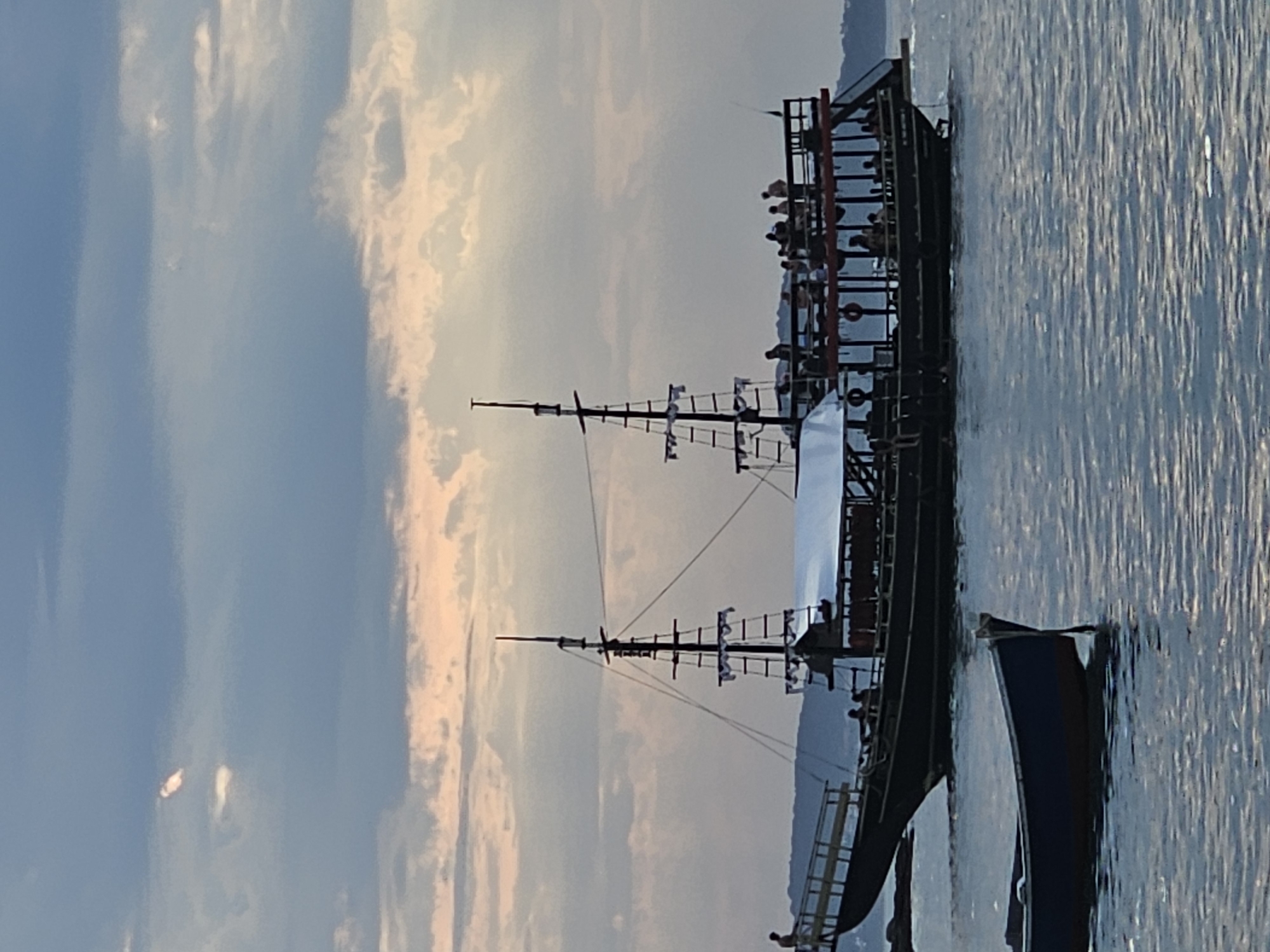 The image captures a large, two-masted ship, possibly a replica pirate ship, sailing on the water off the coast of Armação dos Búzios, Brazil. Several people are visible on the ship. To the right of the ship is another, smaller boat. The sky is filled with clouds.