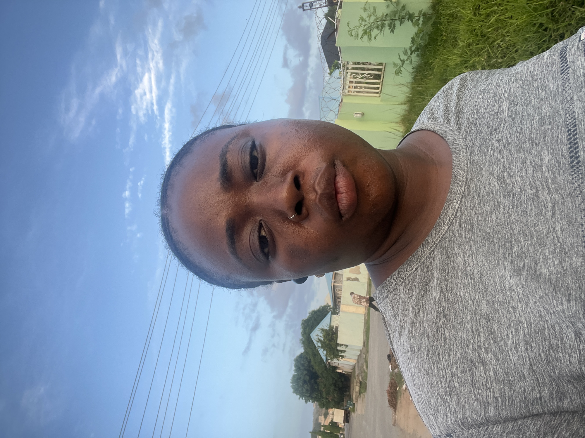 A close-up, slightly angled selfie shows a dark-skinned person wearing a gray heather t-shirt against an outdoor backdrop. The person has a nose ring and appears to be perspiring. Behind the subject, the scene includes a light blue sky with scattered clouds and power lines. In the background are buildings painted in light green, vegetation including grass and trees, and a road. The setting appears to be a residential or suburban area possibly located in Abuja, Nigeria. Other people are visible in the background at a distance.