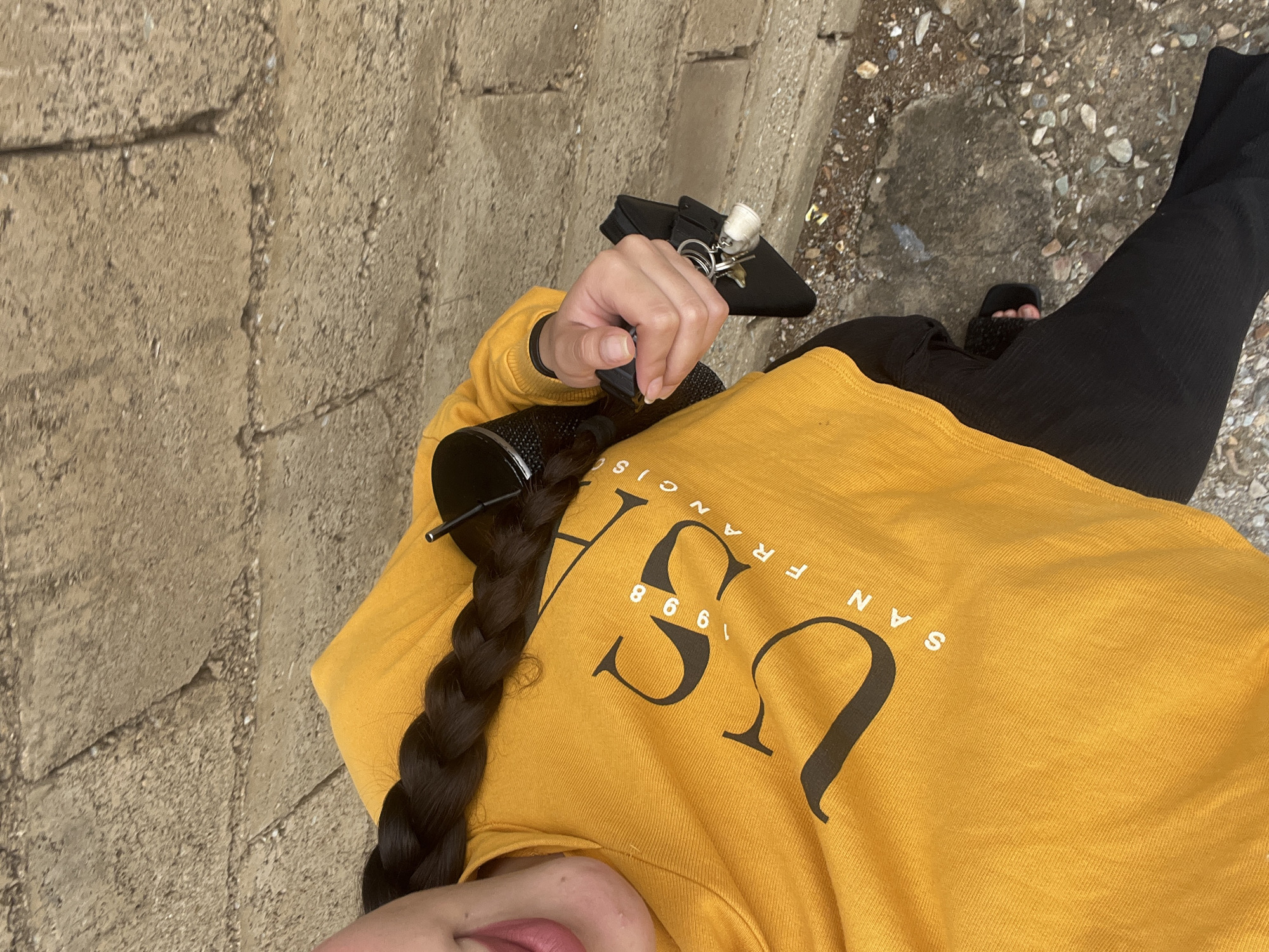 An overhead shot captures a person standing outdoors in Juan Griego, Venezuela, with the frame angled to the right. The individual, largely visible from the upper torso down, wears a mustard yellow long-sleeved sweatshirt and black ribbed pants or leggings. A black furry slide sandal is visible on one foot.
The person has long, dark hair styled in a thick braid that rests over their left shoulder and chest. A black hair clip or accessory is visible in the braided hair. In their right hand, they hold a black water bottle or tumbler and a set of keys on a keyring, which includes a small white decorative charm, attached to a black pouch or wallet.
The ground surface in the image is divided. To the left, there's a light brown concrete block or brick surface. To the right, the ground transitions to a rougher terrain of dirt, small rocks, and gravel, with some wet patches.
Prominently displayed on the yellow sweatshirt is black text: "USA" is large in the center, with "1998" directly beneath it. Arching above "USA" are the words "SAN FRANCISCO". The lighting suggests it is daytime with bright conditions.