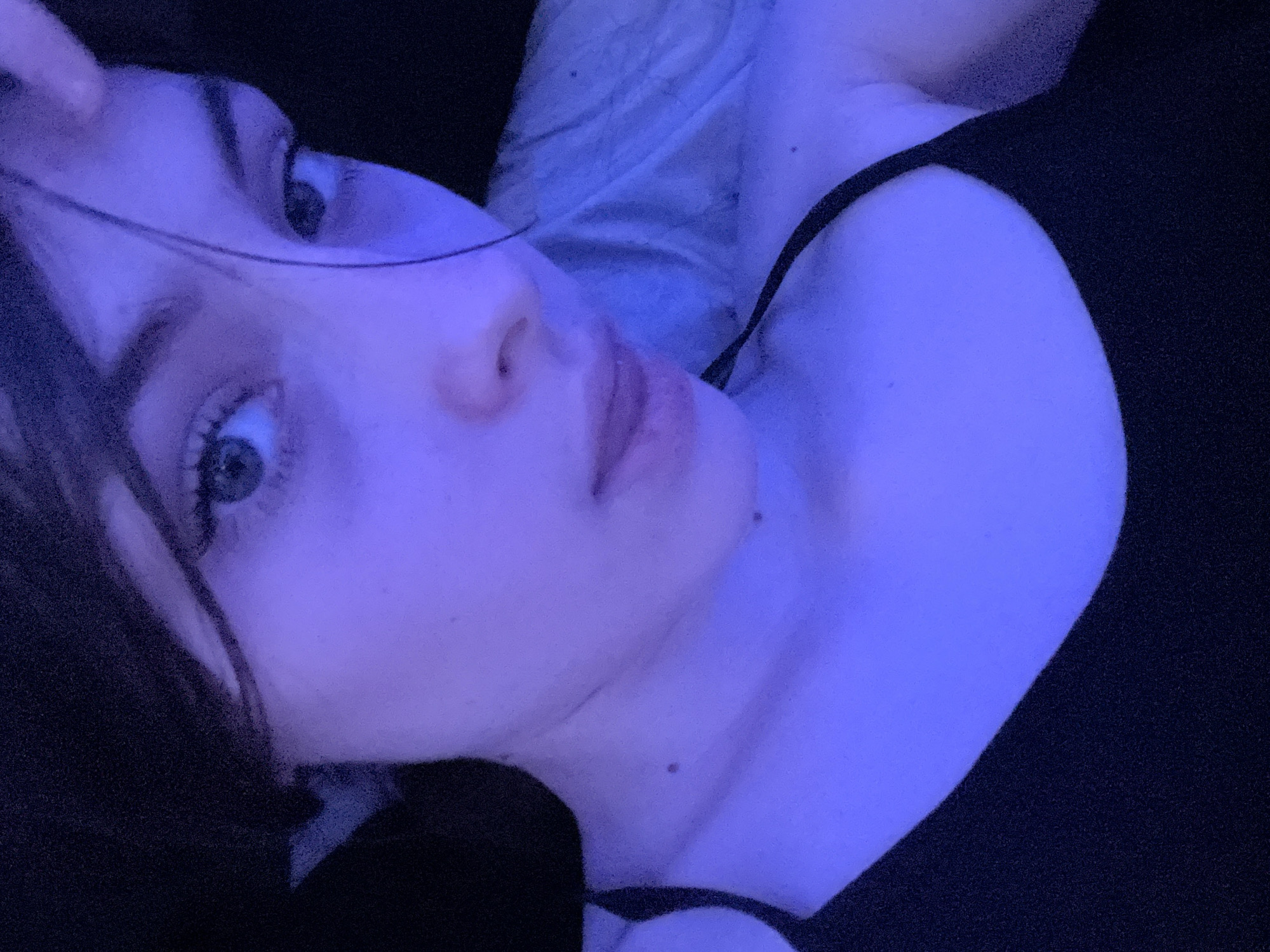 The media file displays a close-up, angled portrait of a young person, likely female, illuminated predominantly by cool blue and purple ambient light. The subject is seen from the upper chest upwards, lying down with their head slightly tilted and looking directly into the camera. Dark hair frames their face, with a few strands falling across their forehead. Their eyes are open and attentive, and their lips are slightly parted. A dark-colored top, possibly a tank top or camisole, with a visible strap over the shoulder, is worn. One hand is partially visible near the top of their head. The background is very dark, almost black, with no discernible objects, placing the sole focus on the individual. The lighting creates a serene and moody atmosphere, suggesting an indoor setting, likely at night or in a dimly lit room. There is no visible text in the image. The implied location context is Sinyavino, Russia.