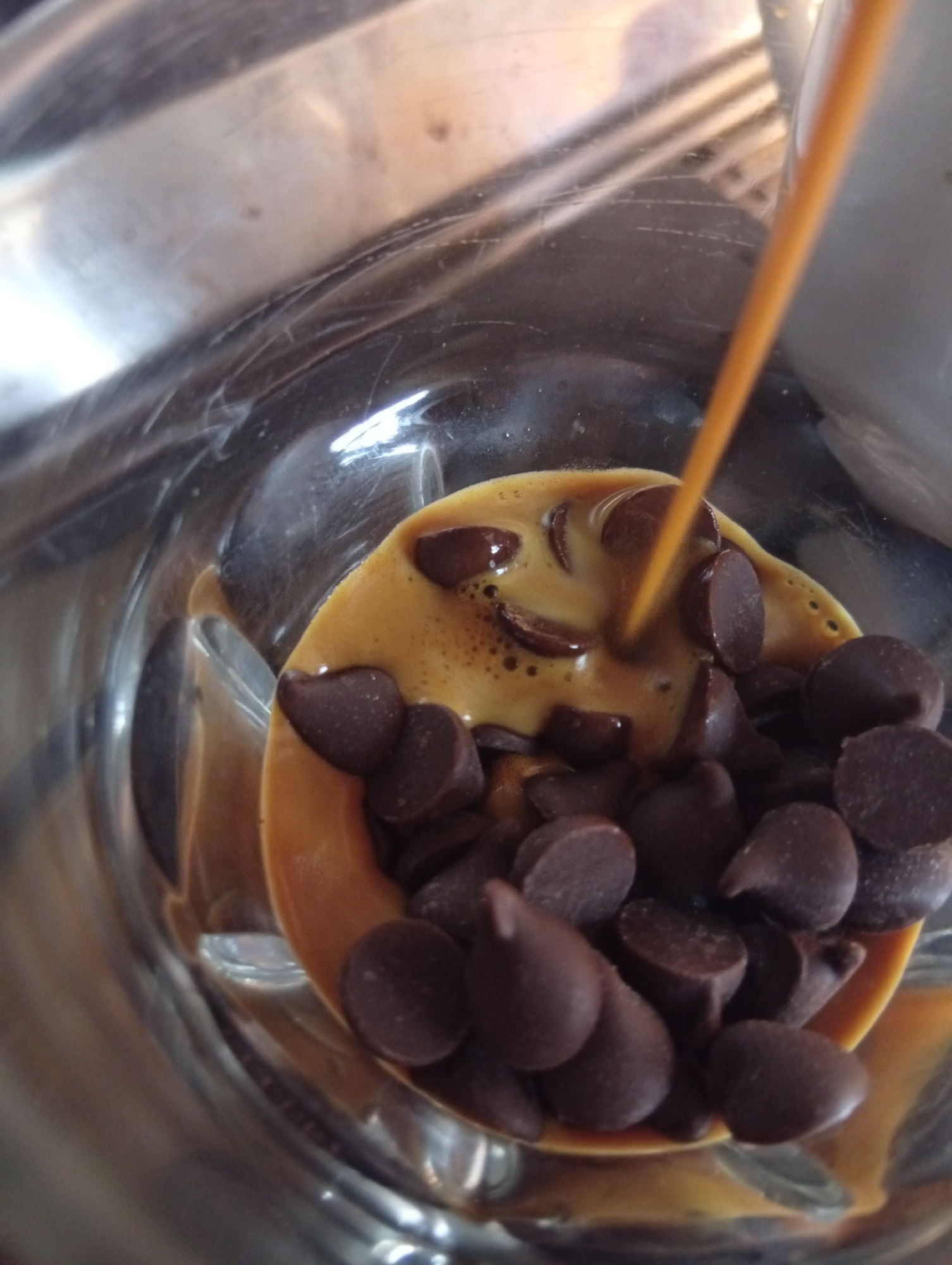 A close-up shot from above depicts chocolate chips in a clear glass container, where a brown liquid is being poured over them. The scene's environment appears to be indoors, possibly in a kitchen or food preparation area. There is no specific indication in the image to confirm a location in Buenos Aires, Argentina.