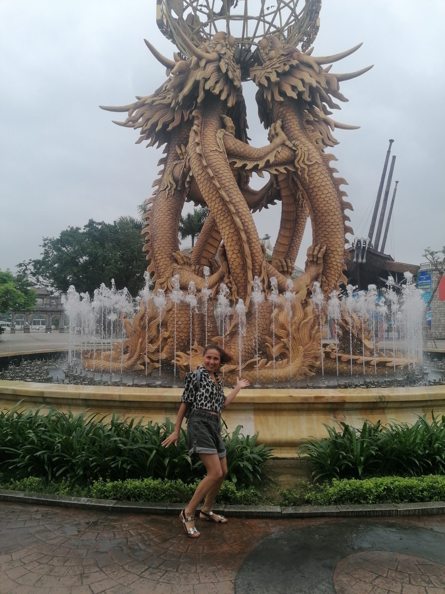 The image captures a woman posing in front of a large, elaborate golden dragon fountain under an overcast sky. The central feature is a multi-headed dragon sculpture, with several golden dragons intertwined, their bodies forming the main structure and supporting a spherical, open-work globe at the top. Water jets vigorously spray from the base of the dragons and around the circular fountain basin, which is lined with green foliage.
In the foreground, slightly to the right, a woman with light brown hair pulled back is smiling at the camera, her hands casually raised. She is wearing a leopard print short-sleeved shirt, dark denim shorts, and embellished sandals. The ground she stands on is a reddish-brown paved surface with a circular pattern, appearing damp.
In the background, to the right of the fountain, a large, dark ship-like structure with multiple masts is visible. To the left, a stone archway or gateway can be seen in the distance, flanked by green trees and other architectural elements. A partially obscured blue sign in the background shows the Vietnamese word "DƯƠNG". The overall setting appears to be a public outdoor space, possibly a park or a themed attraction, in Bai Chay Ward, Vietnam. The time of day is daytime, with muted light due to the cloudy weather.