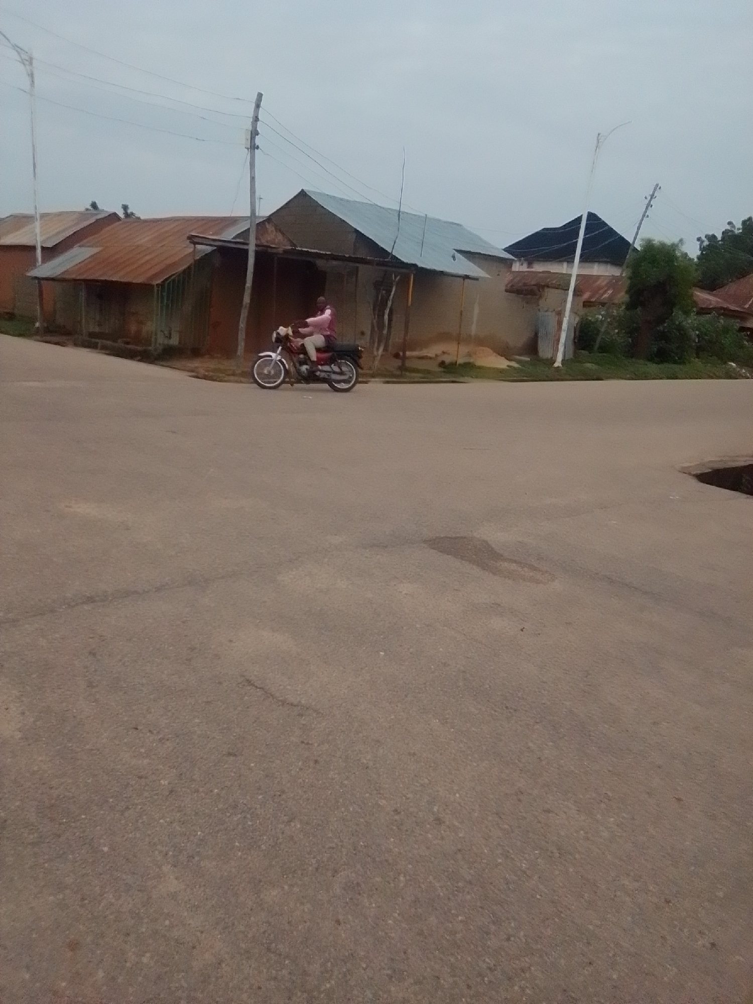 A person is riding a motorcycle on a paved road in Gombe, Nigeria. The motorcycle is red and white, and the rider is wearing a pink shirt and dark pants. The rider is facing forward and appears to be moving. In the background, there are several buildings with corrugated metal roofs. There are also power lines and utility poles visible. The sky is overcast. The overall scene suggests a rural or semi-urban environment.