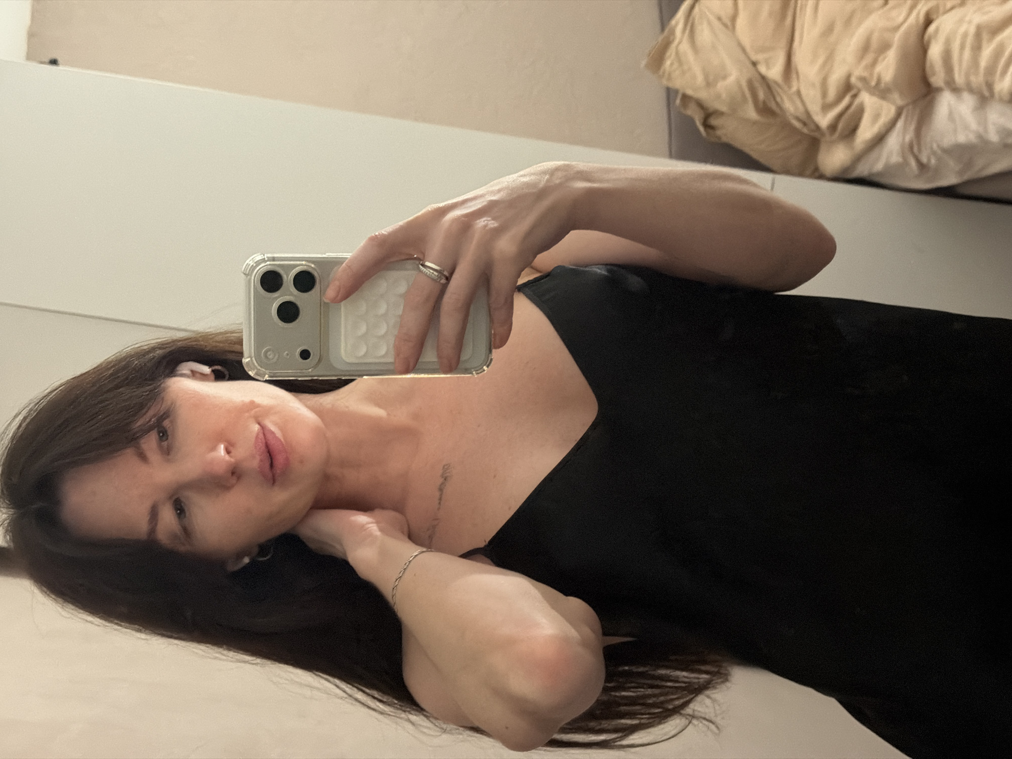 A woman with long, dark brown hair takes a selfie in what appears to be an indoor setting, possibly a bedroom. She's wearing a black top and holding an iPhone with a clear case. A ring is visible on her left ring finger. The background includes a light-colored wall, a closet door and a rumpled beige comforter. There is no evidence within the image to suggest that it was taken in the city of Perm, Russia.