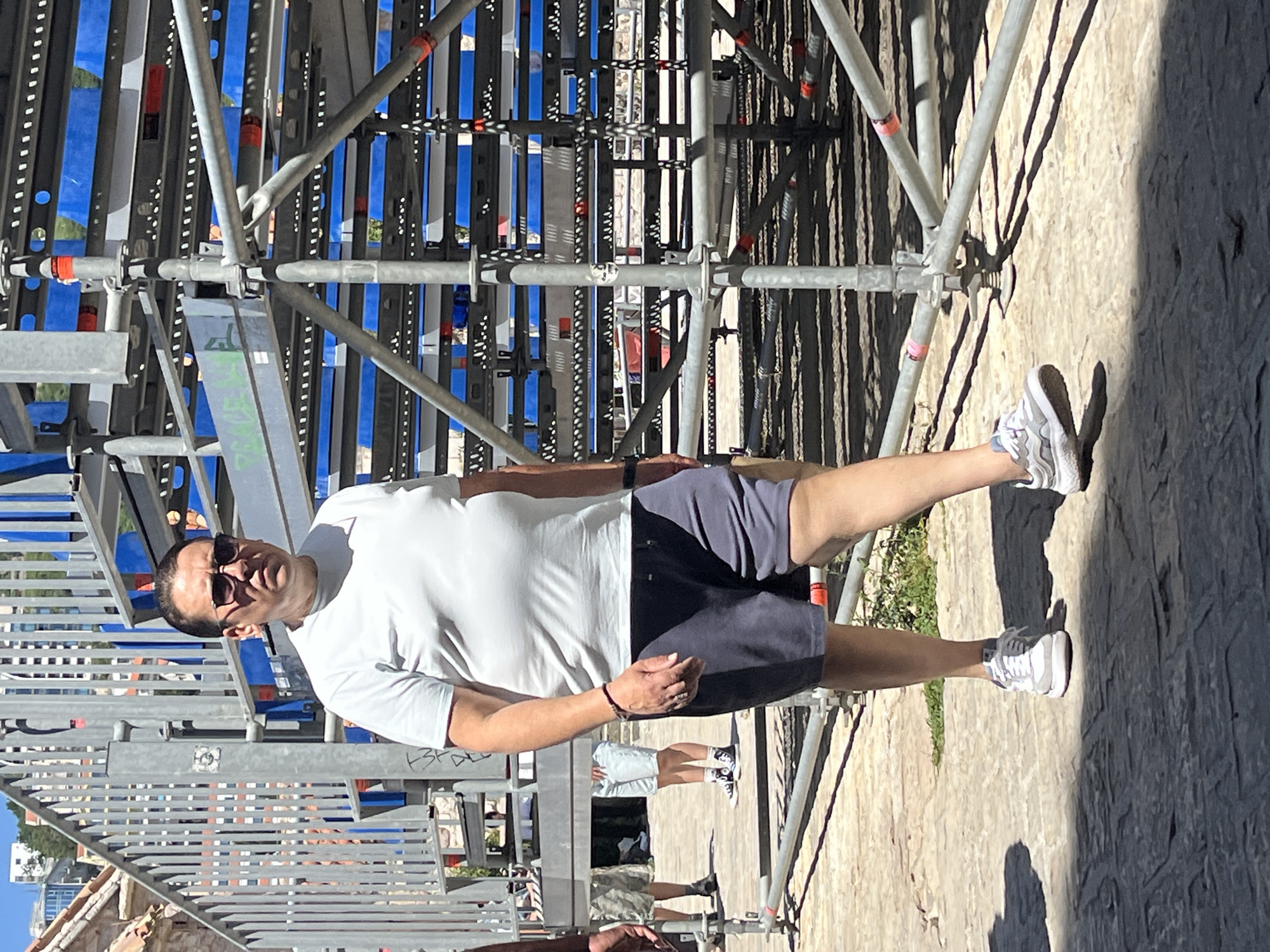 A man wearing a light blue t-shirt, dark blue shorts, and white sneakers is walking on a stone pathway. He is also wearing sunglasses and a bracelet on his left wrist. In the background, there is a metal scaffolding structure and buildings visible. The scene appears to be in an outdoor urban environment, likely in Budva, Montenegro, near the sea given the scaffolding which suggests an event or construction is taking place.