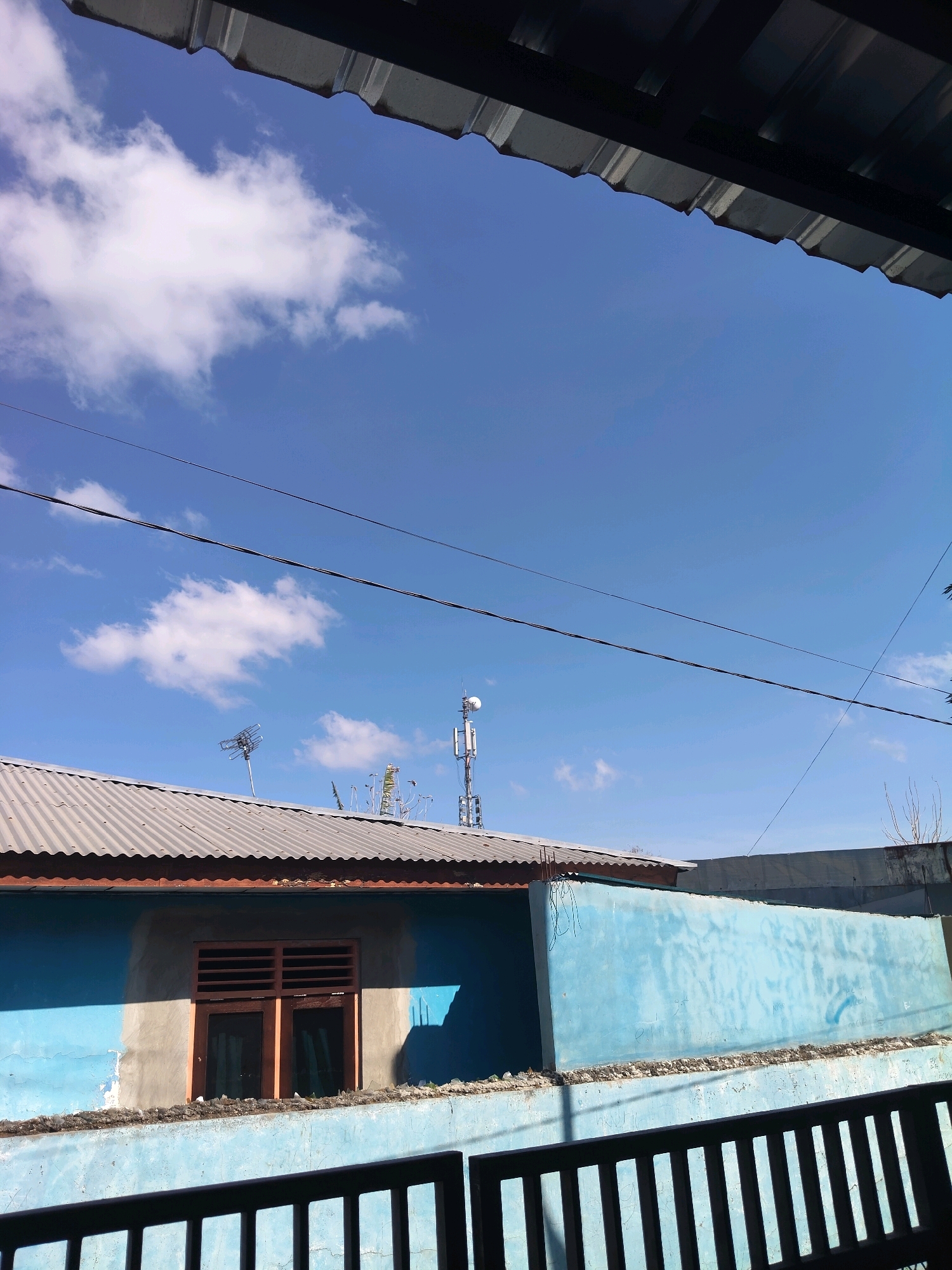This is a vertical, medium-shot, eye-level photograph taken from underneath an overhang, looking out at a bright blue sky with scattered white clouds. The scene depicts the rooftop of a residential building in Kupang, Indonesia. A corrugated metal roof covers the building. A blue painted wall is visible, with a window featuring wooden shutters. Two cell towers and an antenna are prominent structures on the rooftop. Power lines crisscross the sky above the buildings. A dark metal fence is in the foreground, obscuring the lower portion of the scene. The overall impression is one of a clear day in an urban or suburban environment.