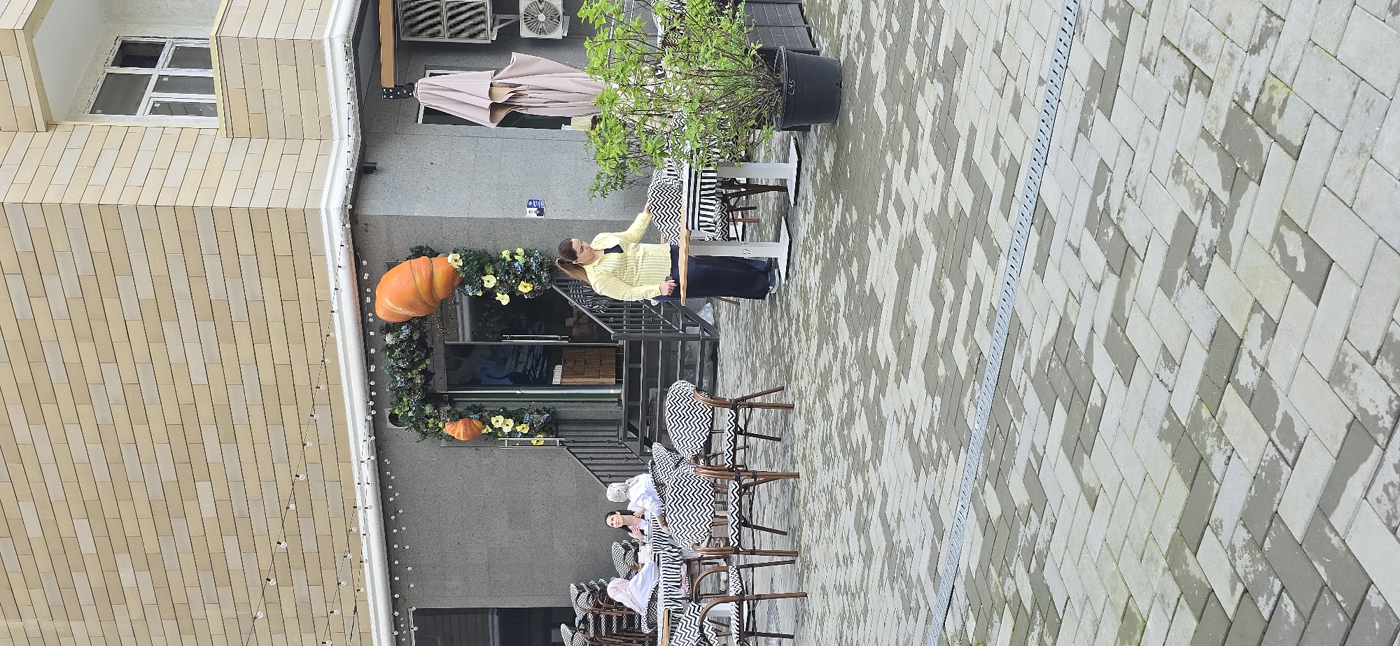 The image captures an outdoor dining area near a building in Grozny, Russia. A brick building with windows is visible on the left side of the frame. A woman with blonde hair is standing next to a staircase in front of a gray building, wearing a yellow shirt and dark pants. Decorations include a garland of greenery, flowers, and oversized pumpkins around the building entrance. A row of tables with chairs, some occupied by people, is set up on a paved area, which shows a pattern of rectangular stones in varying shades of gray. In the background, greenery in a large pot sits next to a table. The scene suggests a restaurant or café preparing for autumn decorations.