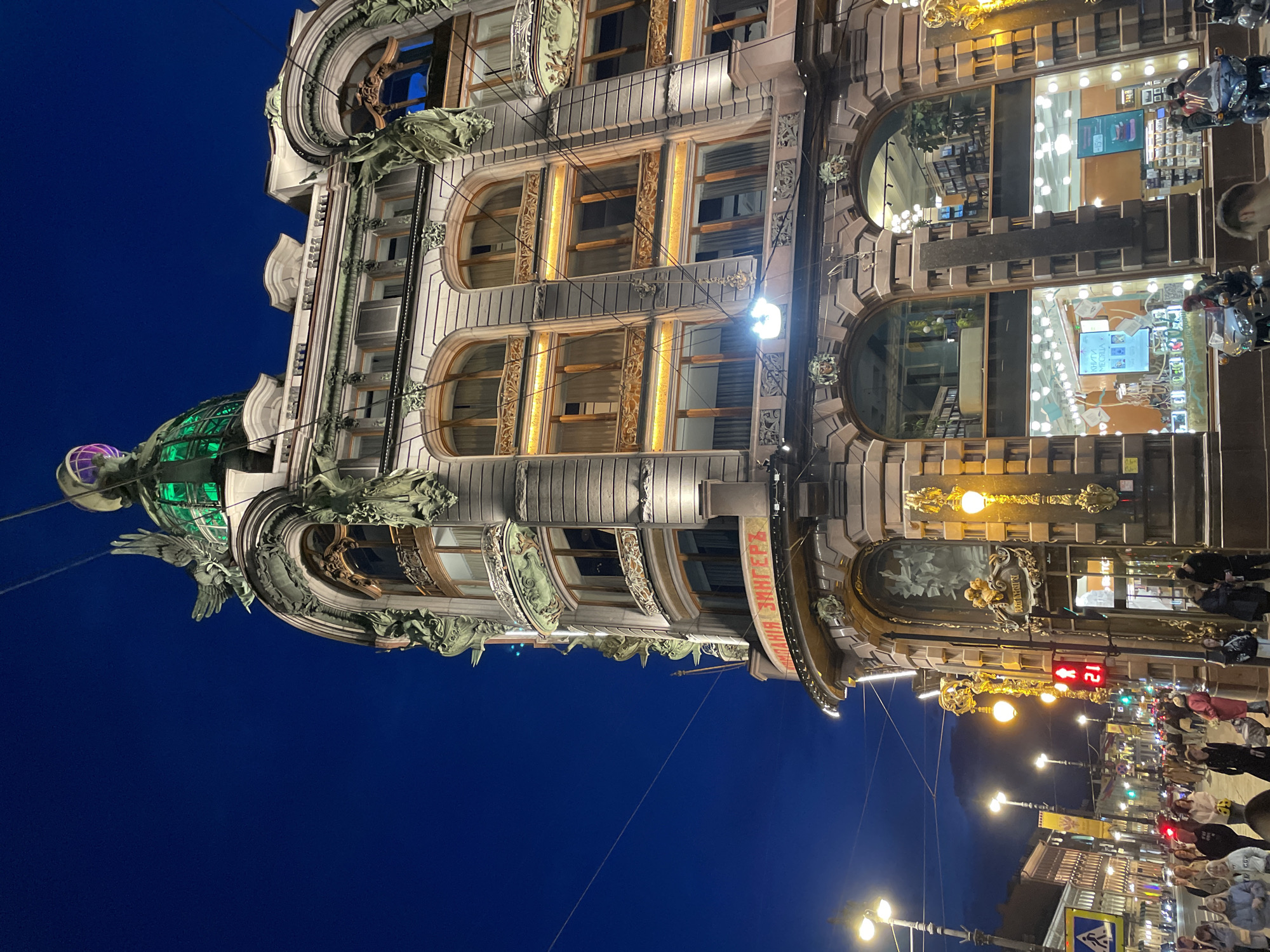 This image, originally oriented 90 degrees clockwise, depicts a grand, multi-story building illuminated at night against a deep blue sky, characteristic of Saint Petersburg, Russia. The architecture is highly ornate, featuring intricate decorative elements, carved statues resembling griffins and human figures, and elaborate window frames. Many windows are arched and glow with warm internal light.
A prominent feature is the large, green, glass dome on the left side of the building, topped with a spherical ornament emitting a soft purple light. The building's facade is primarily light-colored, accented with darker and gilded details.
At street level, a lively urban scene unfolds. Numerous pedestrians are visible on the sidewalk, some gathered near brightly lit shop windows. Streetlights cast pools of illumination, and a red traffic light with the number "21" is seen. The lower floors of the building house businesses with large display windows, one of which shows the text "КНИГИ МЕСЯЦА" (Books of the Month). A curving red banner on the facade clearly displays "ДОМ КНИГИ ЗИНГЕРЪ" (House of Books Singer), identifying the iconic Singer House. Another plaque above an entrance reads "Р.И. ДЕНТР".
The image captures the vibrant atmosphere of an historic city street after dark, highlighting the architectural beauty of the Singer House.