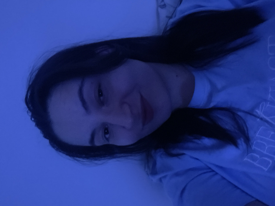 The image depicts a female individual in an indoor setting within the city of Соболиха, Russia. The subject is a young adult female with long, dark hair, a fair complexion, and dark eyes. She is wearing a light blue short-sleeved t-shirt with white text partially visible across the chest, which appears to spell "BREAKFAST" or a similar word. Her lips are colored with a reddish tint, and she is looking directly at the camera with a subtle smile. The entire scene is uniformly illuminated by a strong, cool blue light, suggesting an artificial light source. The background is indistinct, appearing to be a plain interior wall or surface, with a faint suggestion of patterned fabric, possibly a pillow, visible on the far right.
