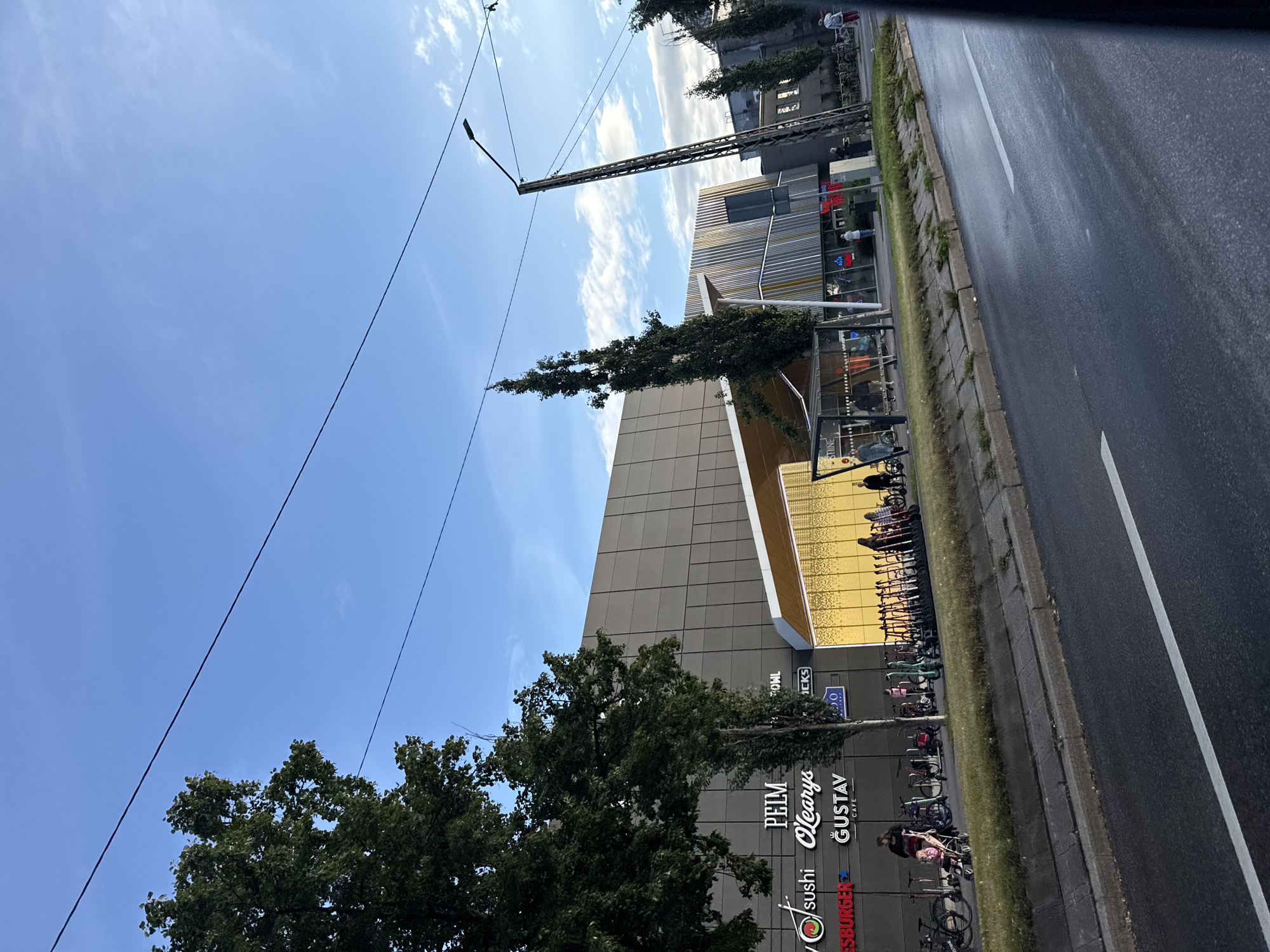 The image captures a street view of a modern building complex in Tallinn, Estonia, likely a shopping center or office building. A row of electric scooters and bicycles are parked along the sidewalk in front of the building. Several trees are strategically planted, with some taller, slender trees flanking the structure. A bus stop shelter is visible, and a few pedestrians are present on the sidewalk. The architecture features a mix of contemporary design elements, including large glass windows and a facade with textured panels. A street lamp stands prominently on the right side of the image. The sky is a clear blue with scattered white clouds, suggesting fair weather. The road in the foreground appears wet, possibly from recent rain. Signs on the building indicate various businesses, including "PEIM," "O'Learys," "GUSTAV CAFE," and "Sushi."