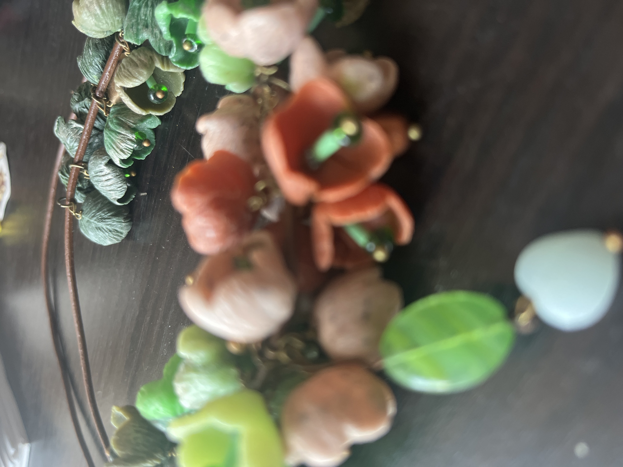 The image displays a close-up, shallow depth of field view of a necklace lying on a dark wooden surface. The necklace features a string of colorful beads that appear to be shaped like flowers and leaves. Some of the flower beads are a peachy-pink hue, while others are a brighter orange. Interspersed among them are green leaf-shaped beads and smaller, light green flower-like beads. In the foreground, a large, translucent green leaf-shaped bead is prominent, followed by a pale blue heart-shaped charm. The necklace itself is suspended from a thin, dark cord. The background is blurred, showing hints of a light-colored object, possibly paper, on the left. The scene is static and focuses on the detail of the jewelry. There are no people or any discernible environmental features of the city of Vladimirovka, Russia, or any other location, visible in the image.
