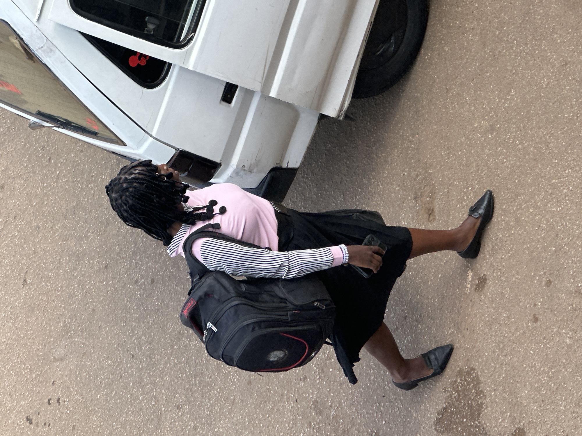 A young African woman with braided hair, wearing a pink and white striped long-sleeved shirt, a black skirt, and black slip-on shoes, walks past the side of a white van in Bukuru, Nigeria. She is carrying a large black backpack with red accents over her shoulders and holds a dark-colored smartphone in her right hand. The ground appears to be asphalt or a similar paved surface with scattered small puddles or stains. The van is white with a slightly damaged rear bumper and a partially open sliding door. Red decorative stickers are visible on the van's windows. The image is taken from a high angle, looking down on the scene.
