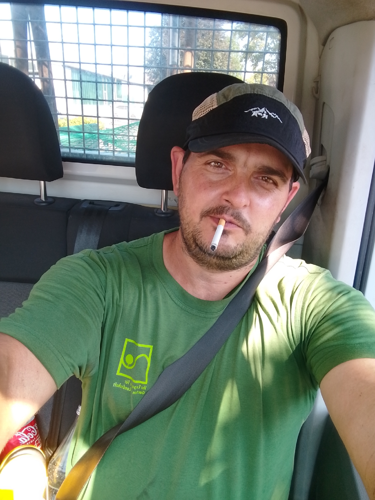 A man with a beard and a baseball cap is sitting in the passenger seat of a vehicle. He is wearing a green t-shirt and a seatbelt. A cigarette is in his mouth. The background shows a window with a metal grate and some greenery outside. The image was taken in Schenefeld, Germany.
