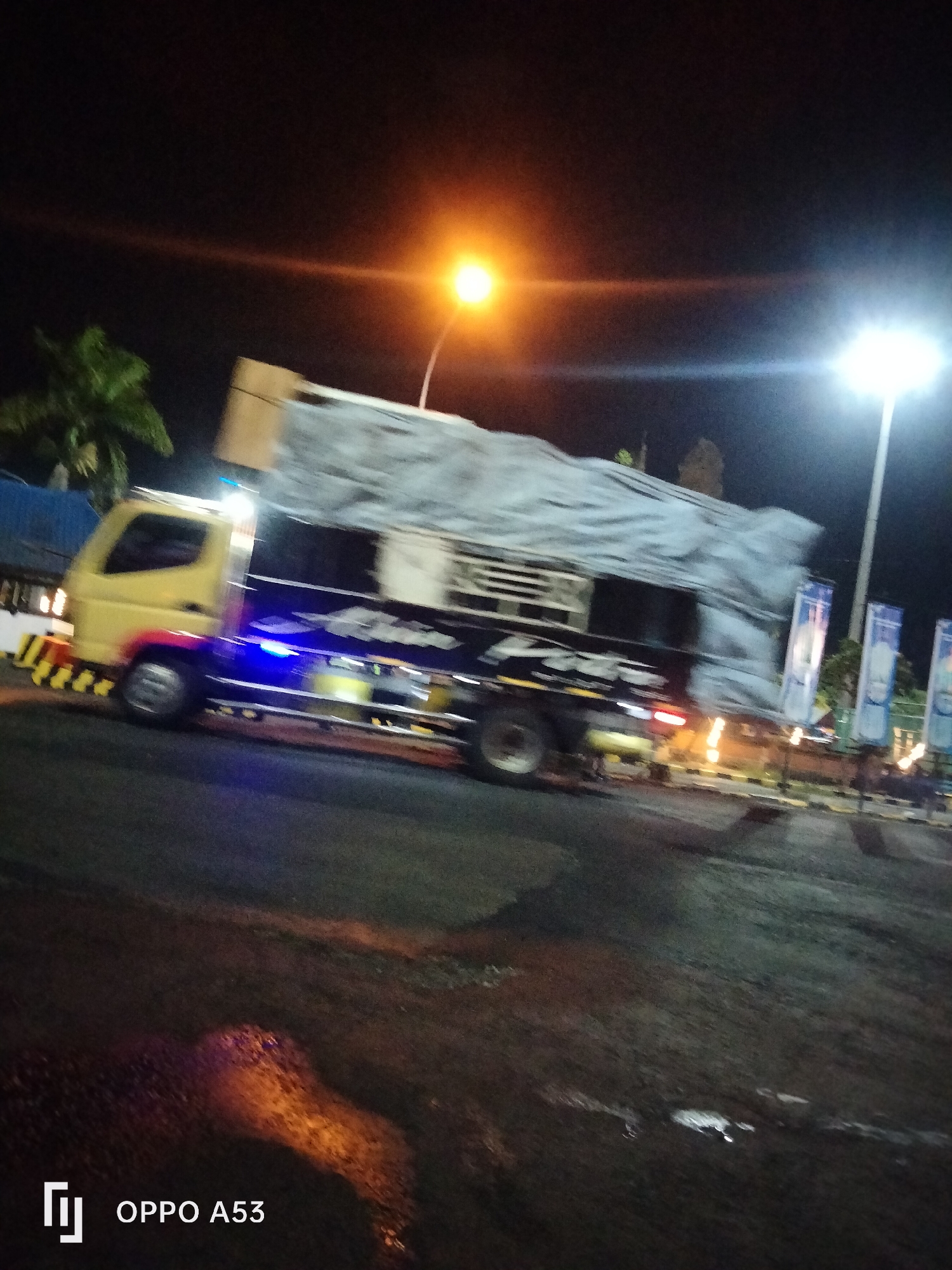 In Melaya, Indonesia, a yellow truck with a gray tarp covers its cargo, driving down a street at night. The truck's headlights and undercarriage lights are illuminated, casting a blue and yellow glow on the road. Yellow and black striped barricades are visible on the side of the road, and two tall banners with blue and white designs stand behind the truck. Palm trees are visible in the background. The ground reflects the streetlights, creating patches of orange and white light. The camera's watermark "OPPO A53" is in the bottom left corner.