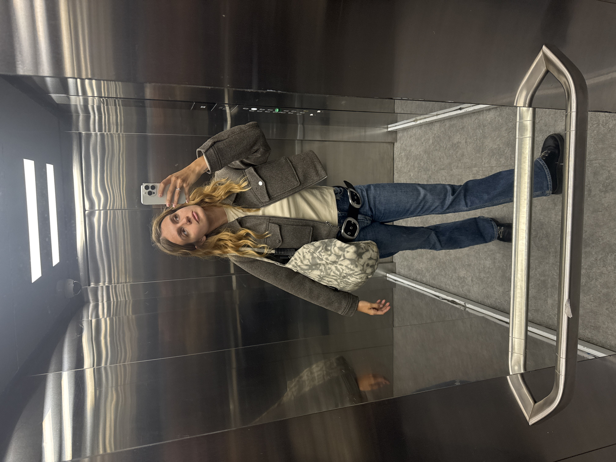 A young woman takes a mirror selfie inside an elevator. She holds her phone in her right hand, showing a silver iPhone with the camera directed at her face. The woman has long blonde hair and wears a beige shirt, a gray jacket, blue jeans with a double-buckle black belt, and black shoes. She also carries a small patterned shoulder bag. The elevator interior is metallic, with a control panel visible at the top, and a handrail to the right. The reflection of the woman and her surroundings can be seen in the mirror. The location is in the city of Unknown, Unknown.