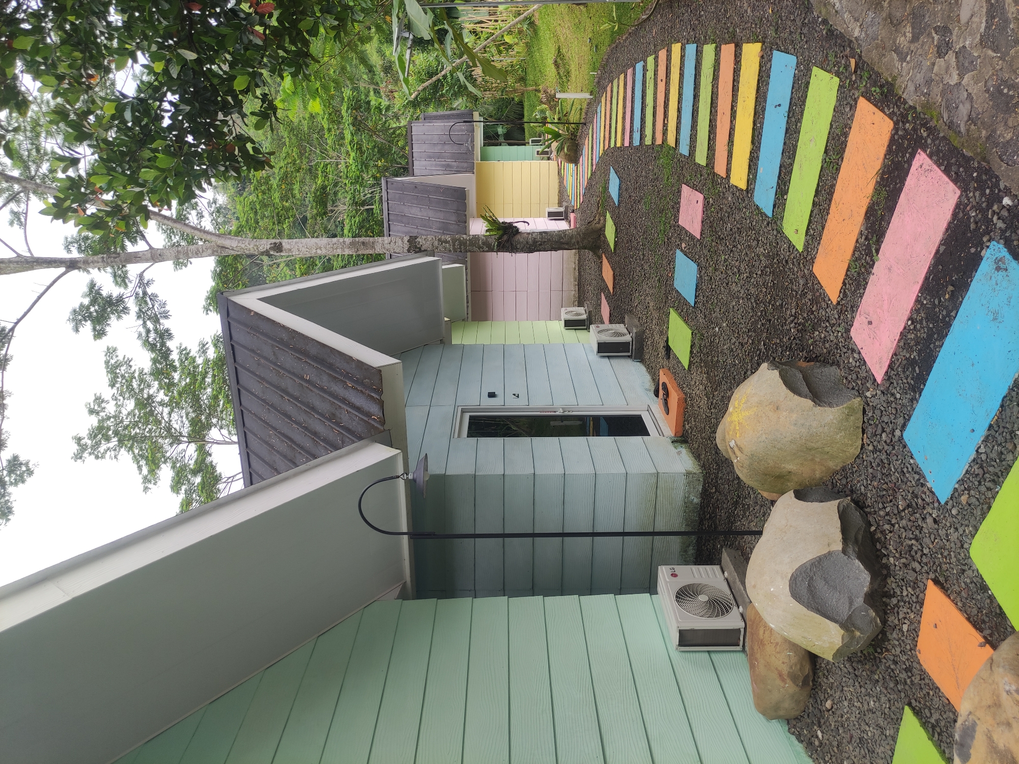 The image captures a row of brightly colored cabins along a pathway in Babakan Madang, Indonesia. The cabins are painted in pastel shades of green, blue, yellow, and pink, with dark grey roofs and light-colored trim. Each cabin has an air conditioning unit mounted on its exterior wall. A path comprised of colorful, rectangular slabs laid over a gravel bed winds between the cabins. Large, irregularly shaped stones are scattered along the path's edge. Trees and lush vegetation surround the structures, suggesting a natural or forested setting. The image is taken from a low angle, emphasizing the vertical lines of the buildings and trees.