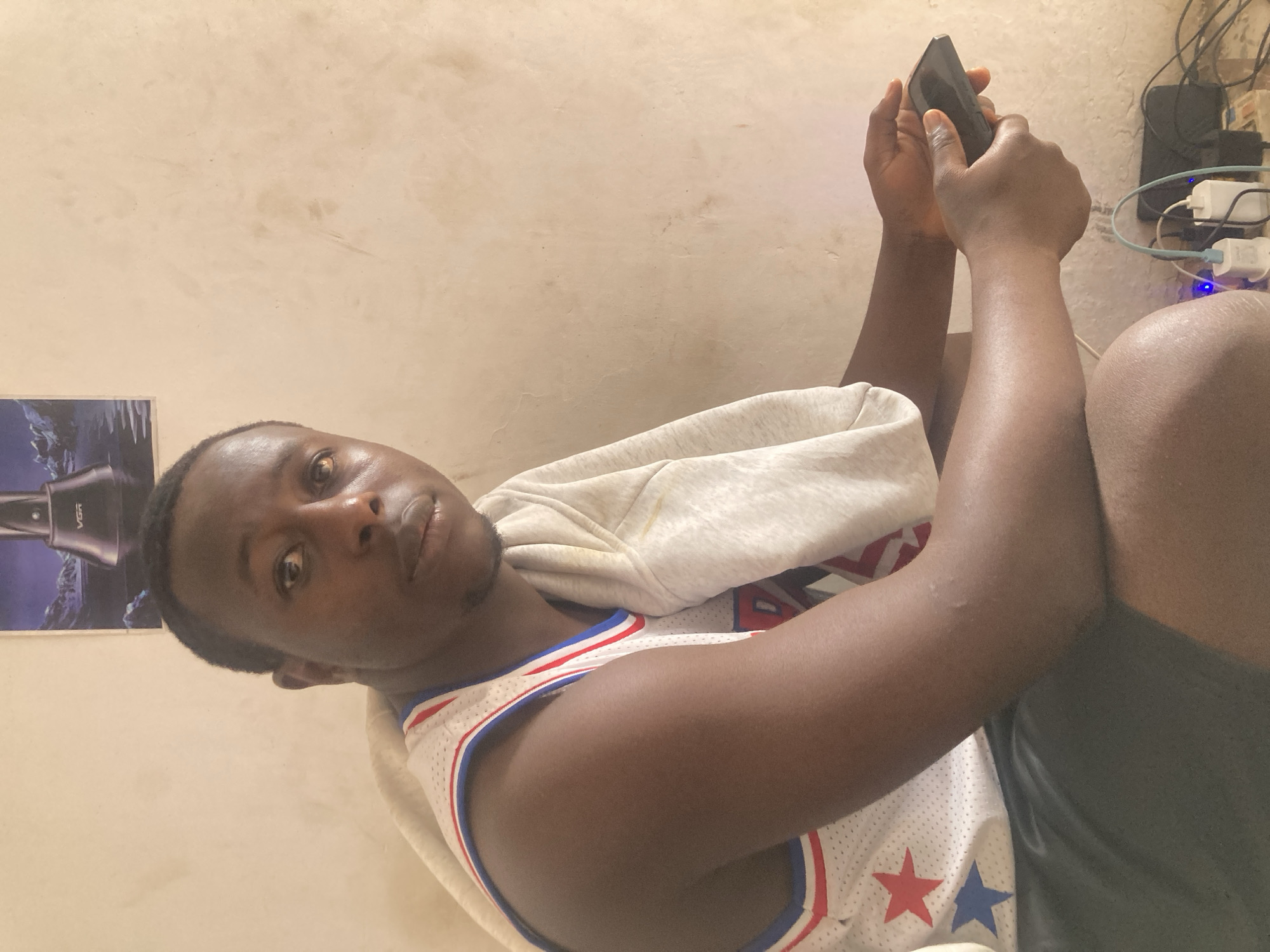 A young African man is lying down indoors, holding a black cell phone in his hands. He is wearing a white basketball jersey with red and blue trim, stars, and numbers, and a light grey hooded sweatshirt draped over his shoulders. He is also wearing dark-colored shorts. A power strip with multiple plugs and cords is visible on the wall next to him, along with a poster. The location is Gwong, Nigeria.