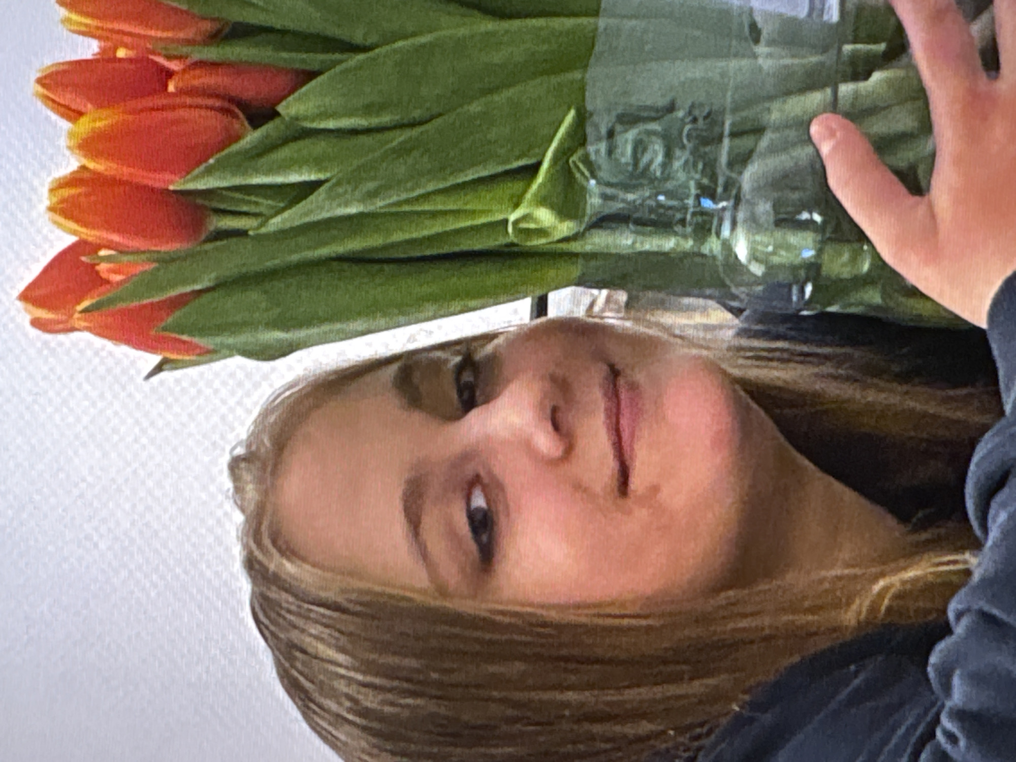 A close-up, medium shot shows a young female with blonde hair and light skin, facing the camera and holding a vase of red and orange tulips. Her left cheek is closer to the camera, and she is looking slightly to her right. Her hair is parted on the left and falls past her shoulders. She is wearing a dark blue hooded sweatshirt. The vase, made of clear glass, is partially visible on the right side of the frame and contains water. The background is a textured white wall. The image appears to have been taken indoors. There is no information in the image to determine its location in Warsaw, Poland.