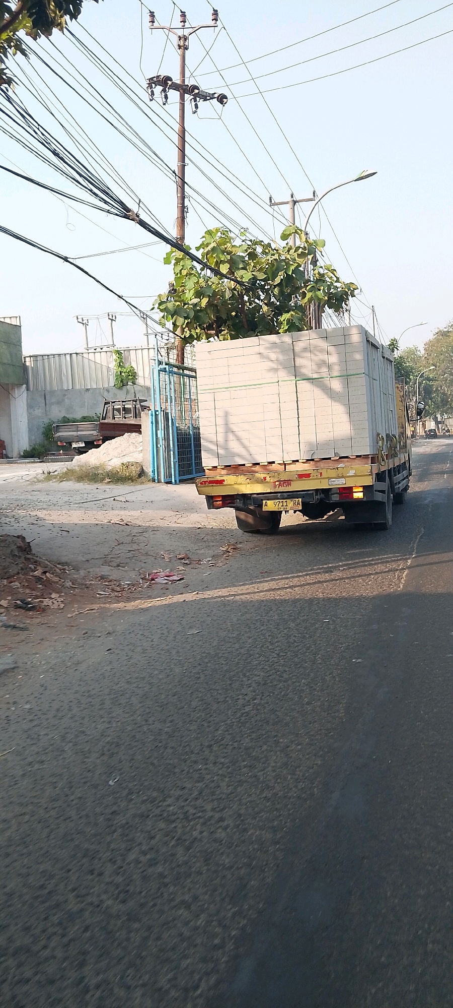 An outdoor, daylight, eye-level, medium shot captures a yellow truck carrying a large load of white bricks. The truck is on a paved road with a rough, dirt shoulder. In the background, a factory with a corrugated metal wall is visible, along with a blue metal gate and some trees. Power lines crisscross the sky. The license plate on the truck reads "A 9711 RA". The scene appears to be in Bekasi, Indonesia, a city known for its industrial areas.
