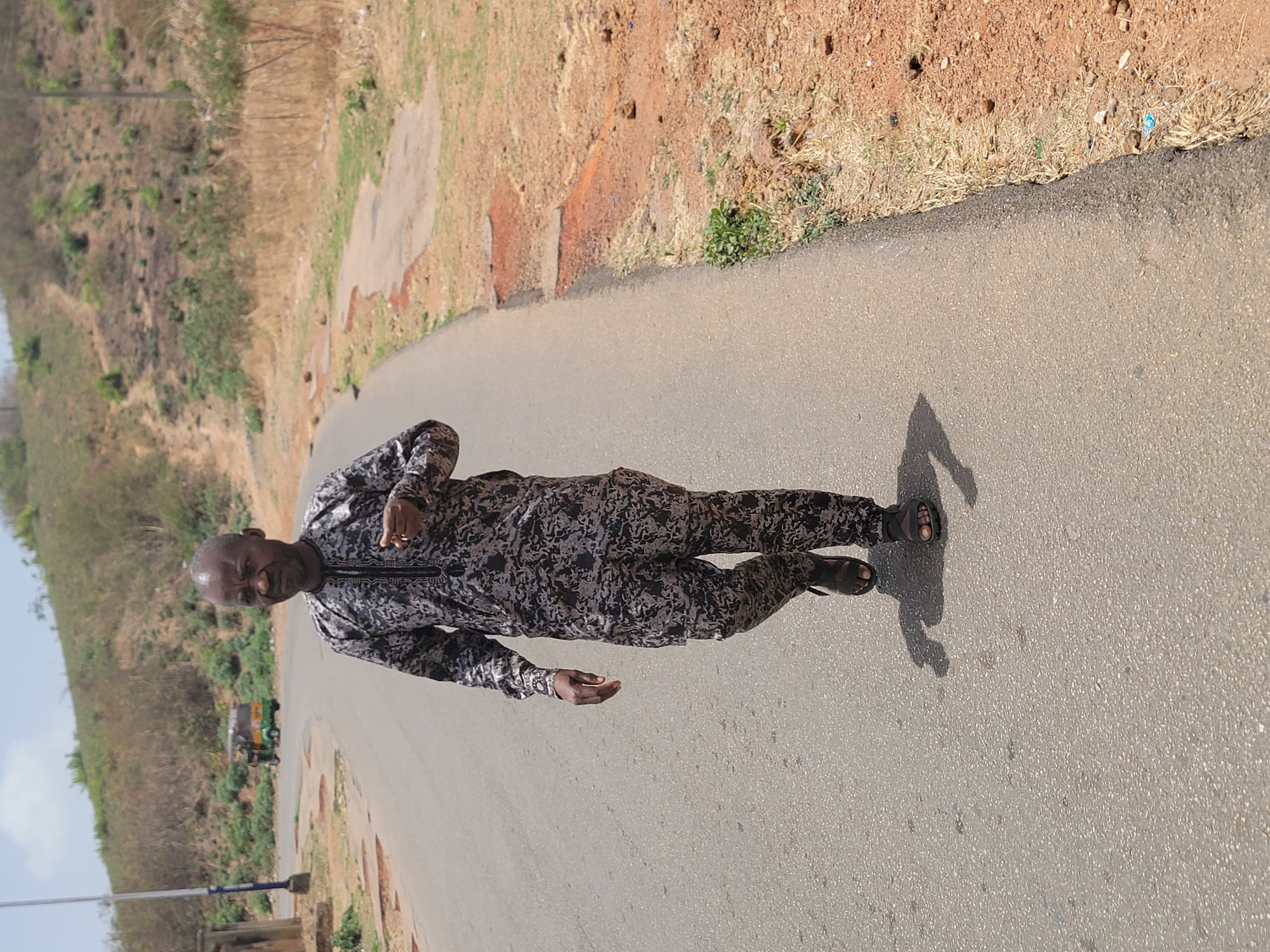 A person is standing on a paved road. The individual is wearing a patterned long-sleeved shirt and pants, and sandals. A shadow is cast on the road next to the person. To the left of the road, there is a hillside with vegetation. On the right, the ground is covered with dry grass and some small patches of green vegetation. In the background, a vehicle can be seen parked on the left side of the road. The scene appears to be in a rural part of Jos, Nigeria.